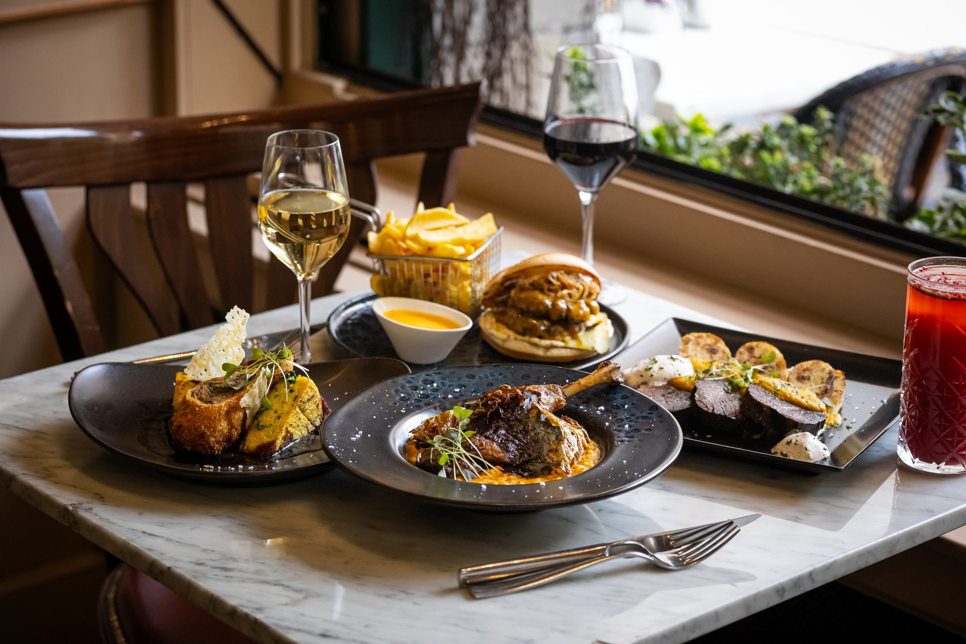Lunch plates on a marble table by the window at Centrál Kávéház: a roast duck leg on barley risotto, a smashed burger with fries and cheddar sauce, chicken Wellington with a parmesan crisp, sliced beef with bread dumplings, and a glass each of white and red wine