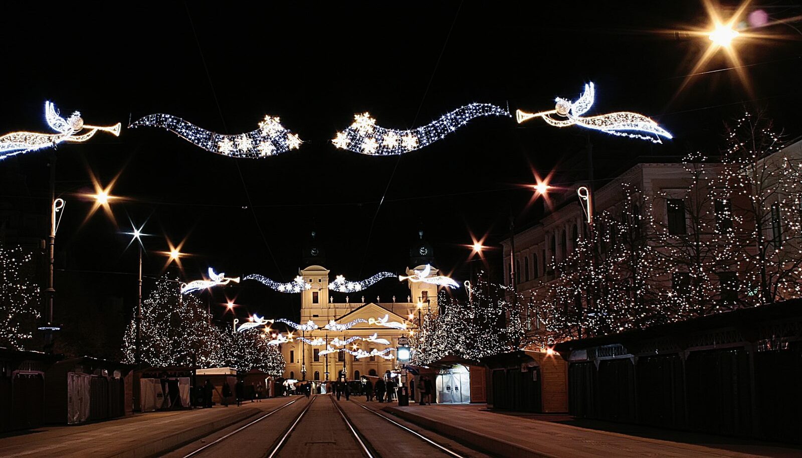 Piac utca à Debrecen la nuit, avec des guirlandes lumineuses d’anges et d’étoiles au-dessus des rails du tram, des arbres enveloppés de guirlandes et des rangées de chalets de l’Avent menant aux deux tours du Grand Temple réformé