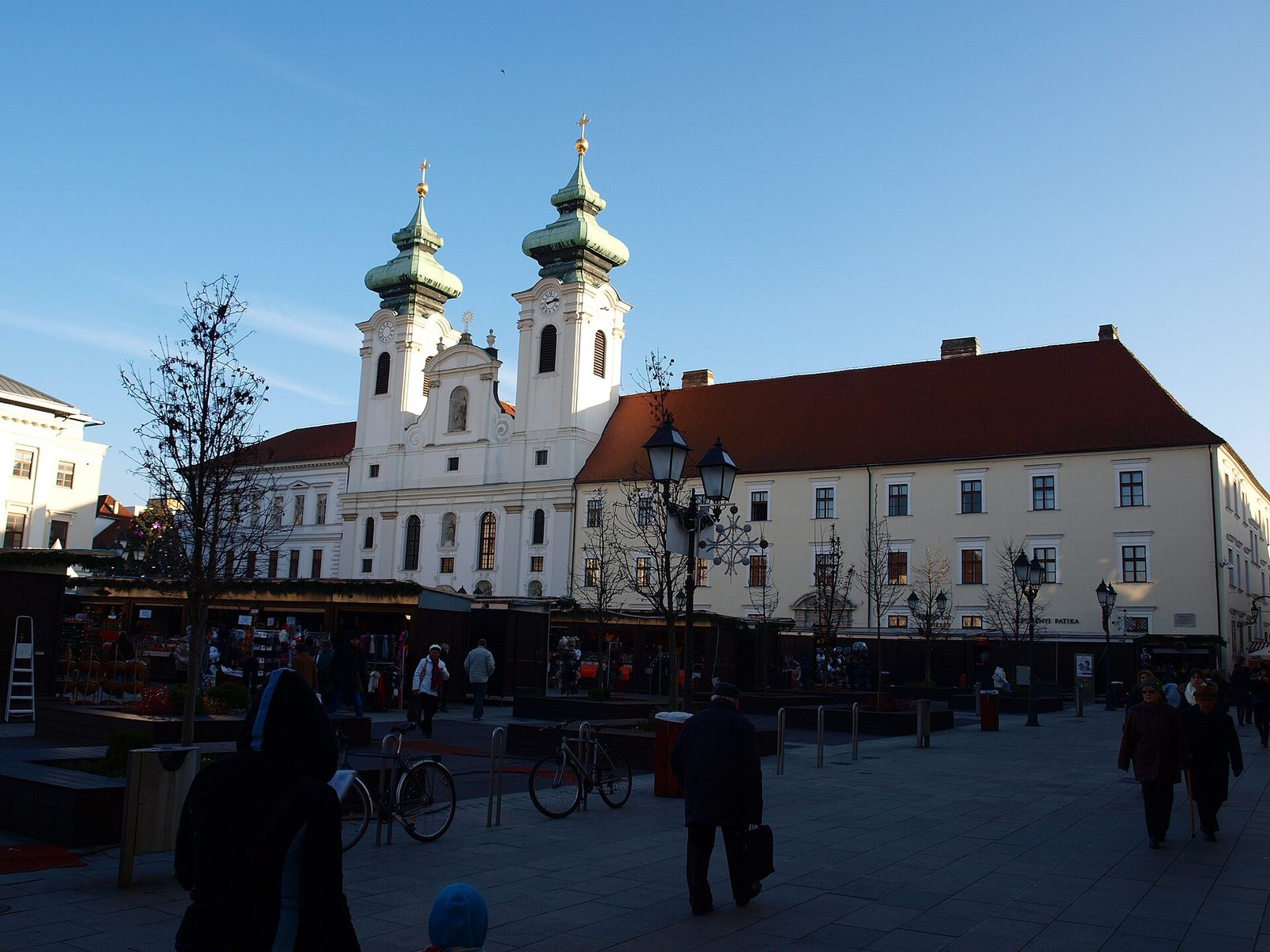 Les chalets du marché de l’Avent sur Széchenyi tér à Győr par un après-midi ensoleillé de décembre, devant l’église blanche Saint-Ignace et ses deux tours, avec un flocon lumineux sur un lampadaire
