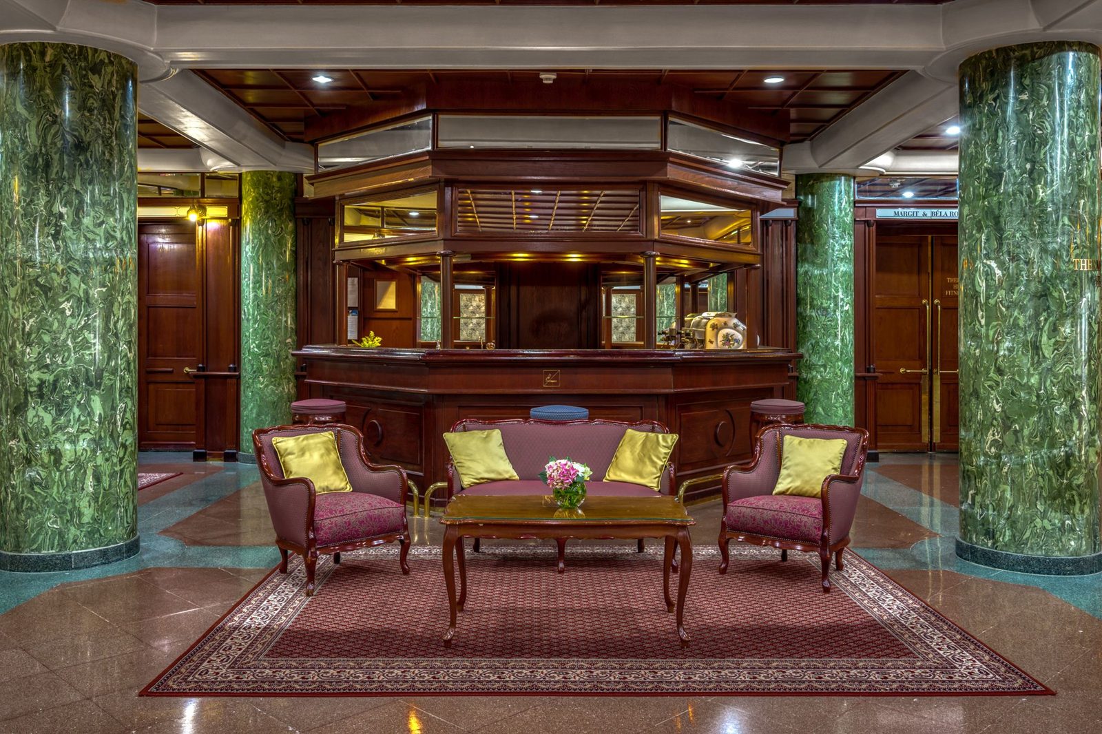 The lobby of the Ensana Grand Margaret Island, with green marble columns, a wooden reception desk and red armchairs