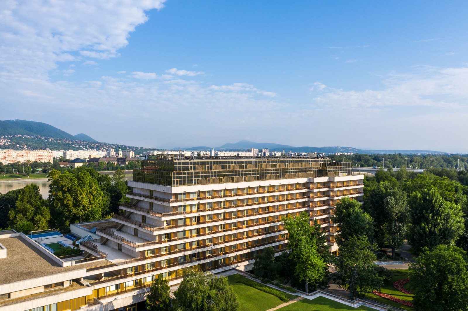 The Ensana Thermal Margaret Island from the air, a long terraced hotel block among the trees, with the Danube and the Buda hills behind