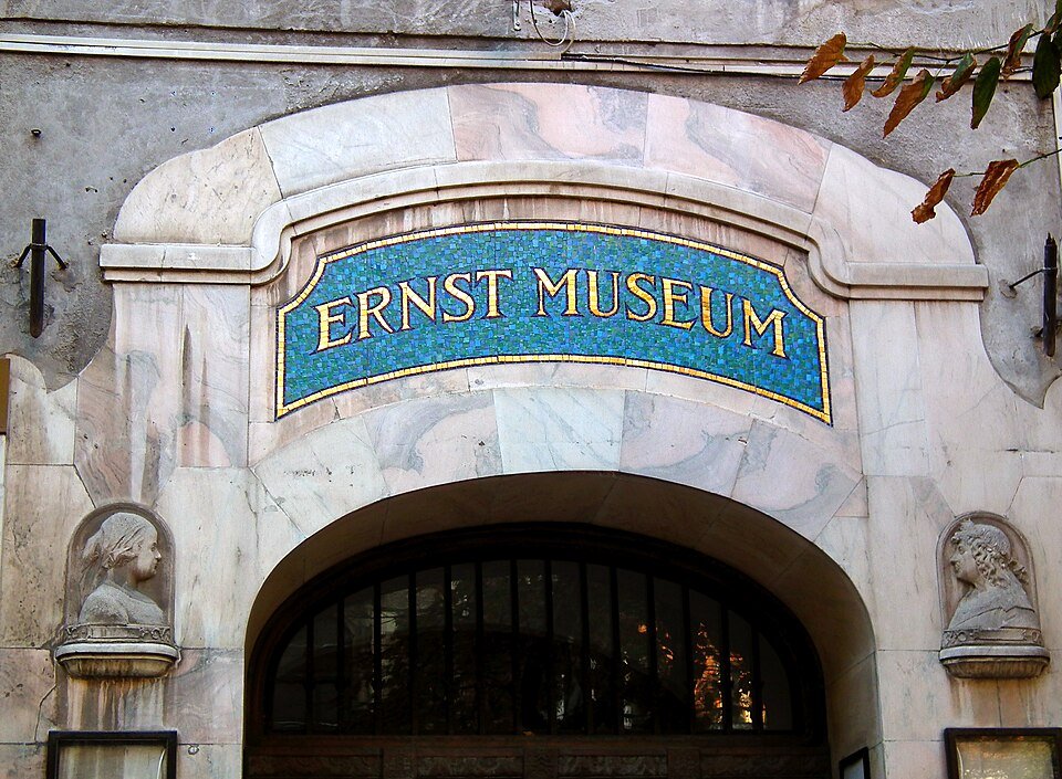 Modrozlatý mozaikový nápis Ernst Museum nad vchodem do Nagymező utca 8, po obou stranách reliéfní busta ve výklenku