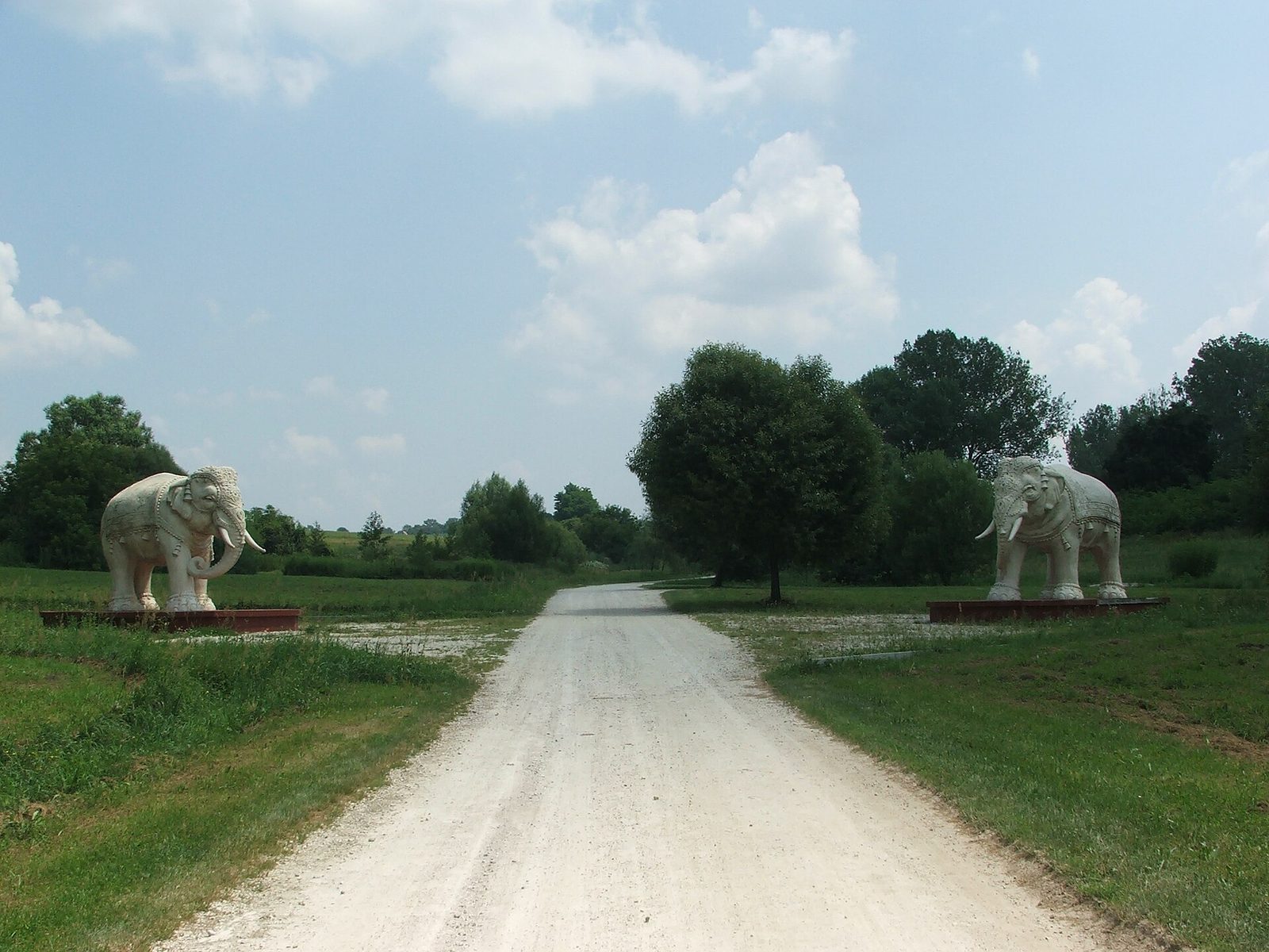 Two carved stone elephants on either side of a white gravel road leading into the green fields of Krishna Valley at Somogyvámos