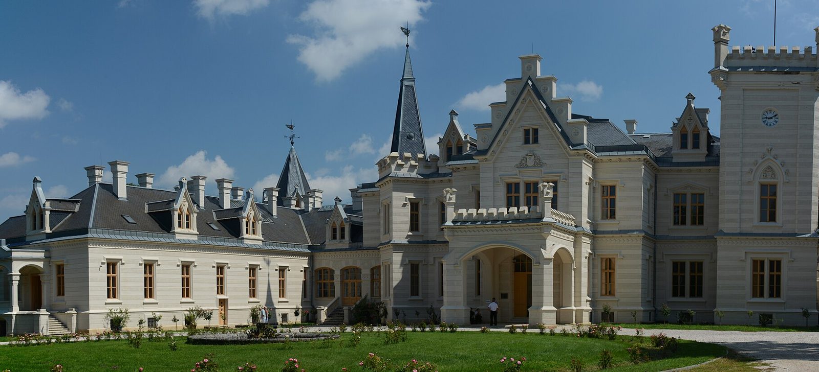 The garden front of Nádasdy Castle, a cream neo-Gothic manor with battlemented towers, steep slate roofs, pointed turrets and tall chimneys above a lawn with flower beds