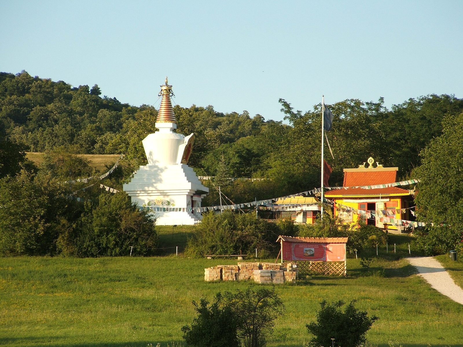A white Tibetan stupa with a gilded spire on a green meadow at Tar, with prayer flags strung to a colorful shrine house and wooded hills behind