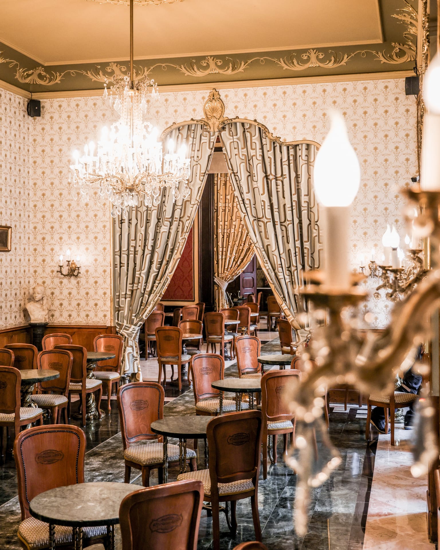 Una sala del Café Gerbeaud con un lampadario di cristallo, tende drappeggiate attorno a un passaggio e sedie di legno ai tavolini tondi di marmo