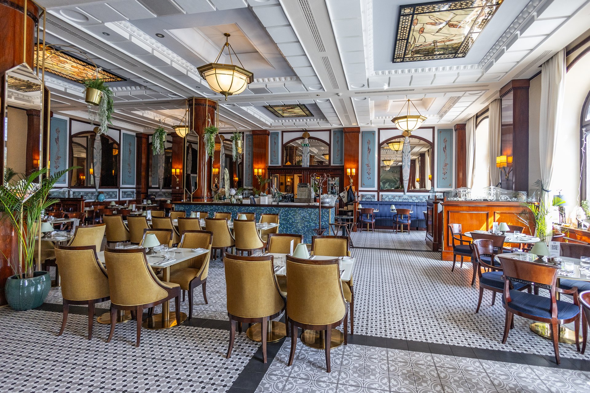 Gundel’s day dining room with mustard velvet armchairs, stained-glass ceiling panels, brass lamps and patterned floor tiles