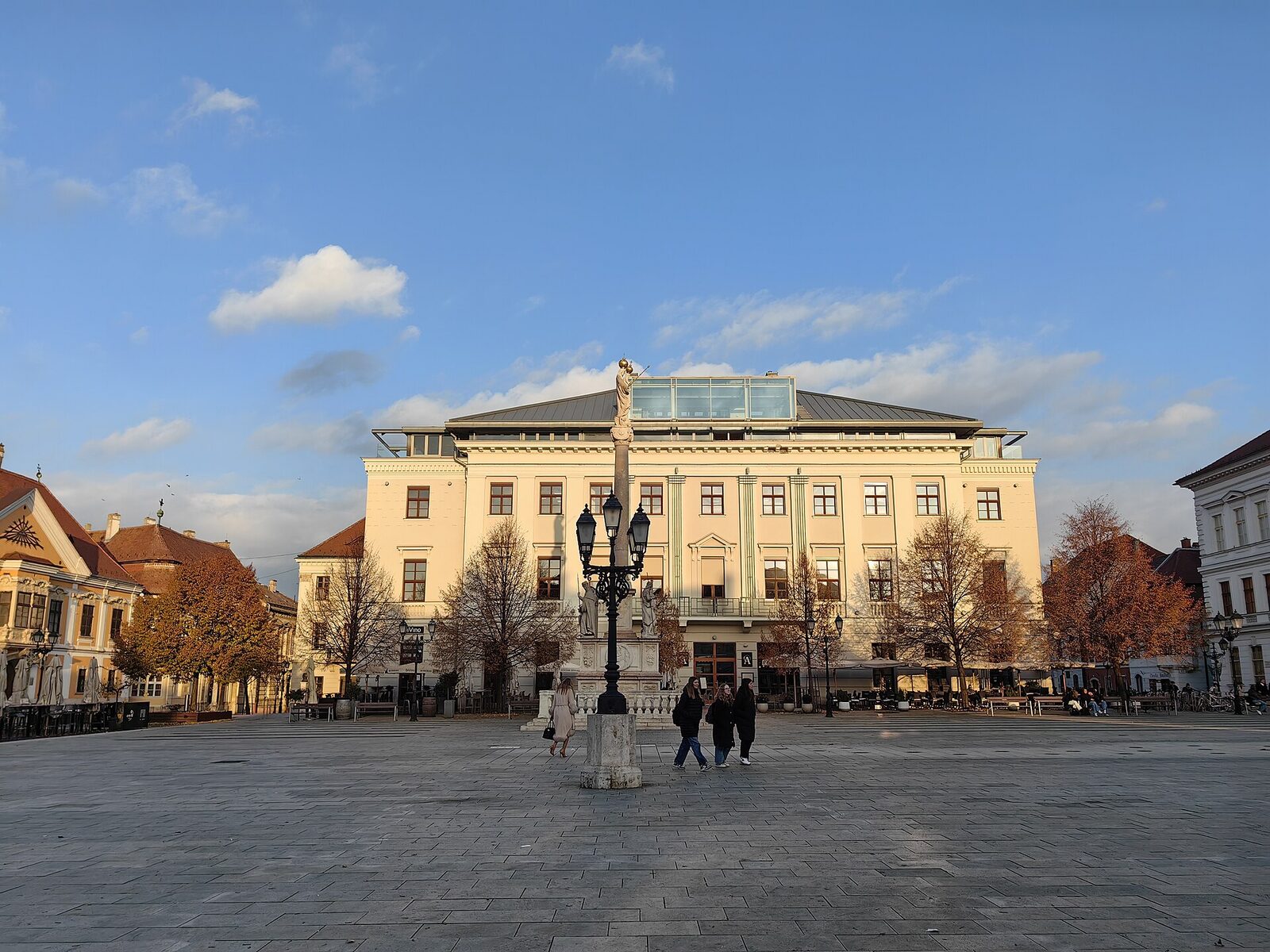 Széchenyi tér in Győr on a sunny autumn day, with baroque houses, a column with a statue on top and trees turning yellow