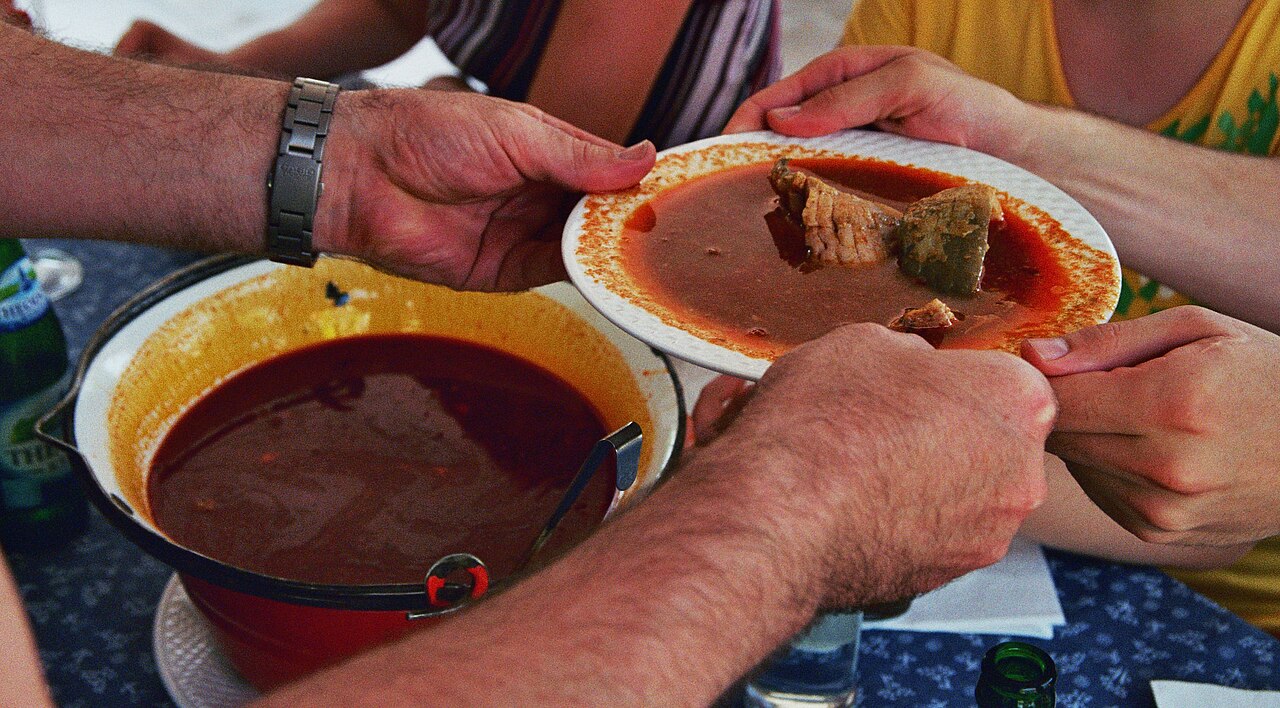 A plate of red halászlé with pieces of fish being passed across a table next to the pot it was served from, in Szeged