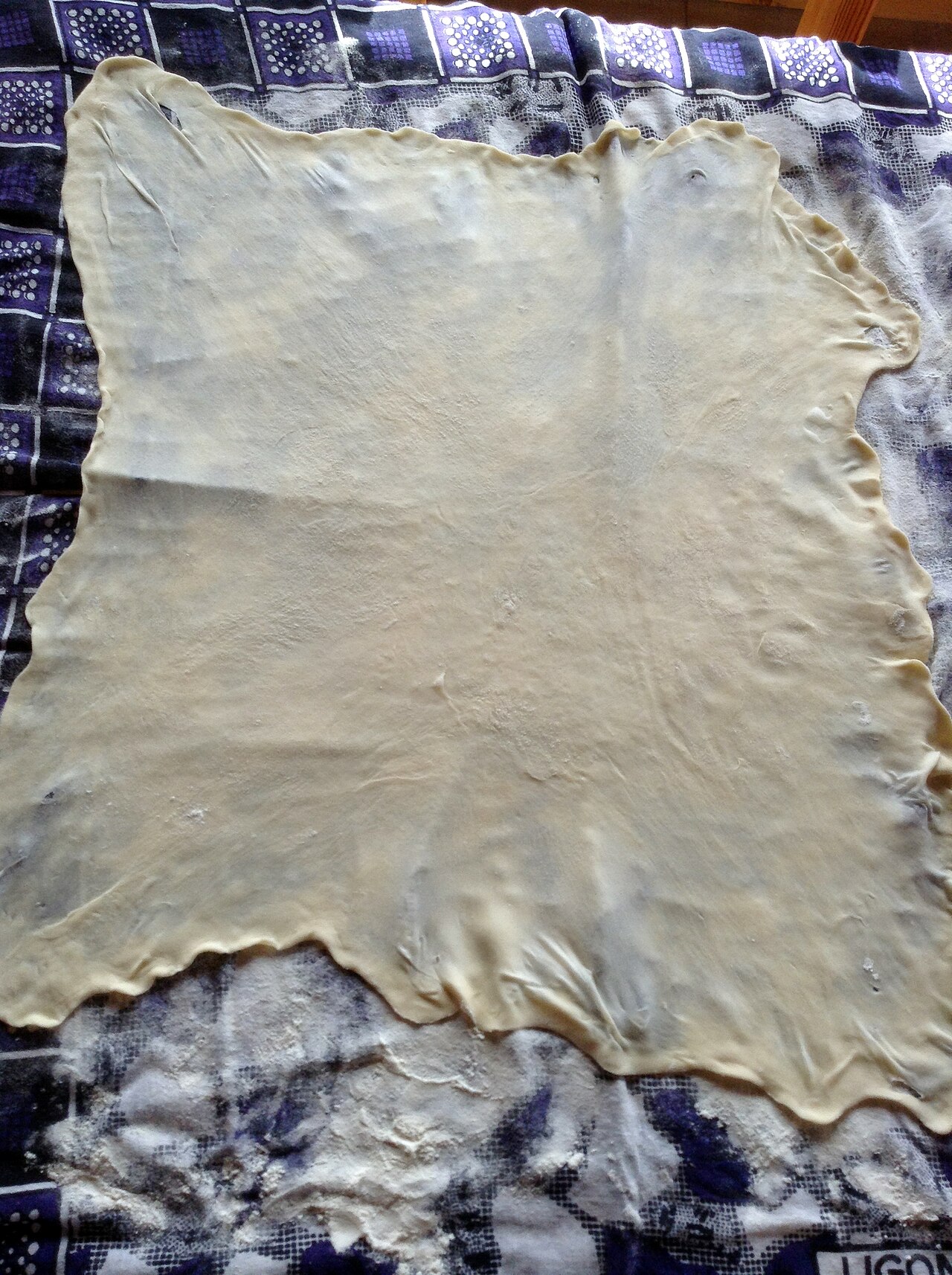 A sheet of dough stretched paper-thin and translucent over a floured patterned cloth on a table