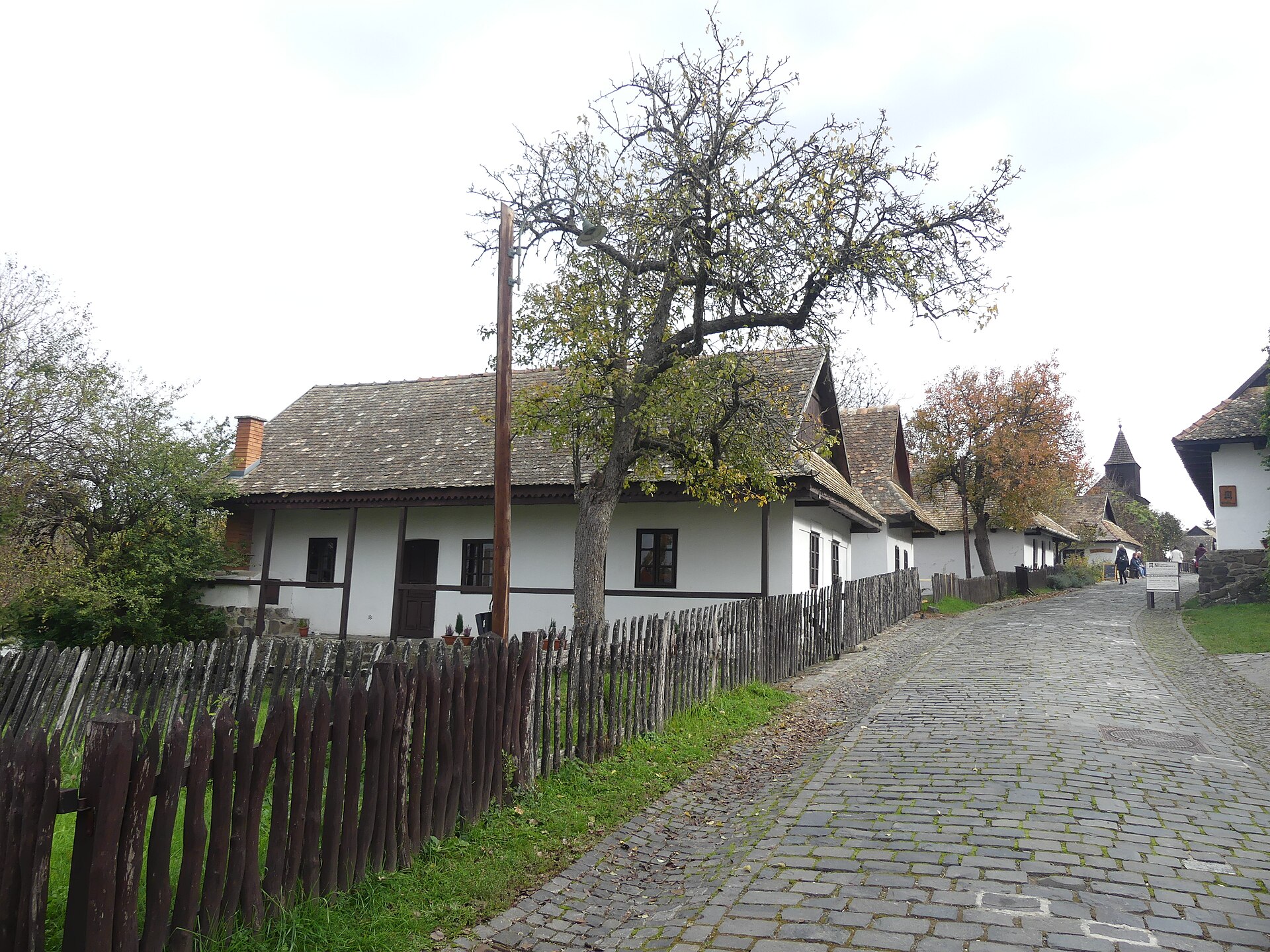A cobbled street in Hollókő lined with whitewashed Palóc houses, with the church tower at the far end