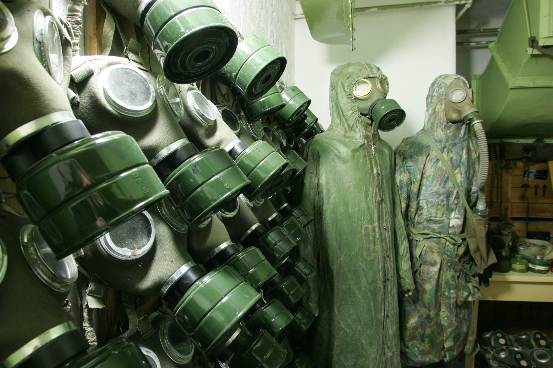 Rows of old gas masks with green filter canisters hanging on a bunker wall in the Hospital in the Rock, next to two figures in rubberised protective capes and gas masks