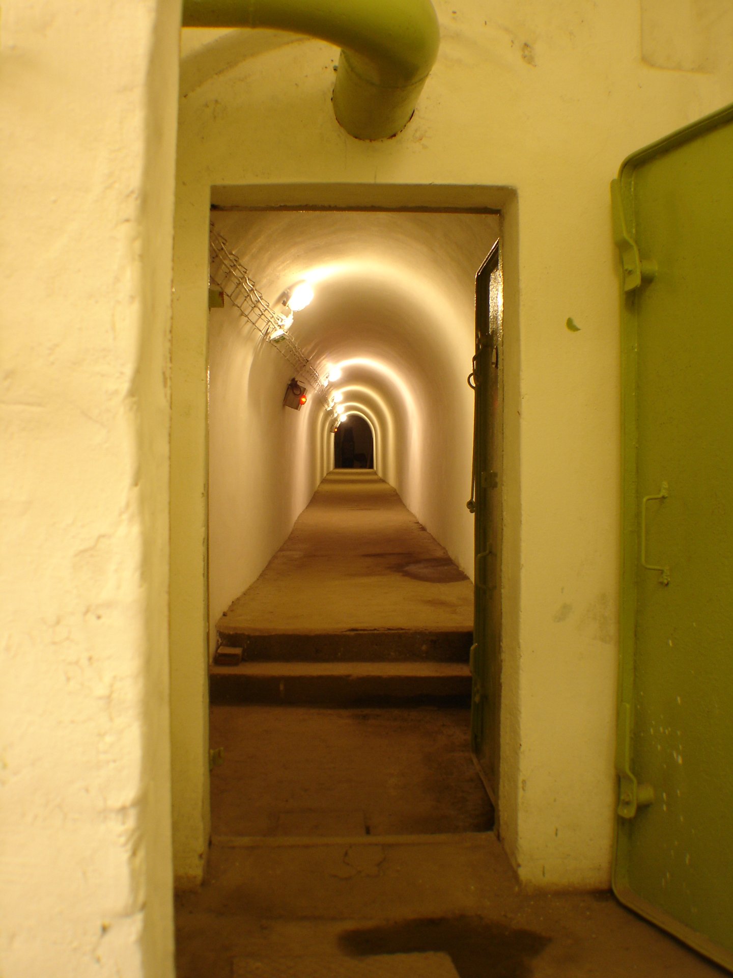 A narrow whitewashed tunnel in the Hospital in the Rock, lit by wall lamps, seen through a doorway beside an open green steel door