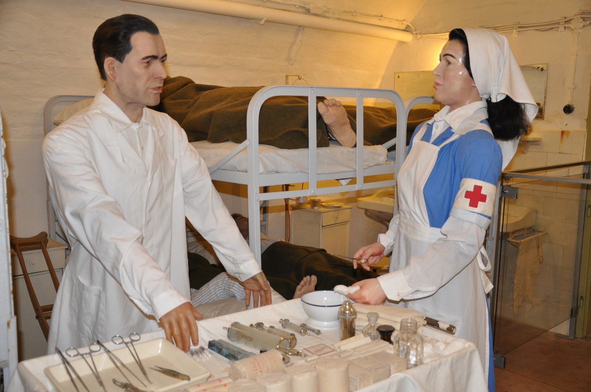 Wax figures of a doctor in a white coat and a nurse with a Red Cross armband beside a trolley of surgical instruments, with patients in metal bunk beds behind them, in a ward of the Hospital in the Rock