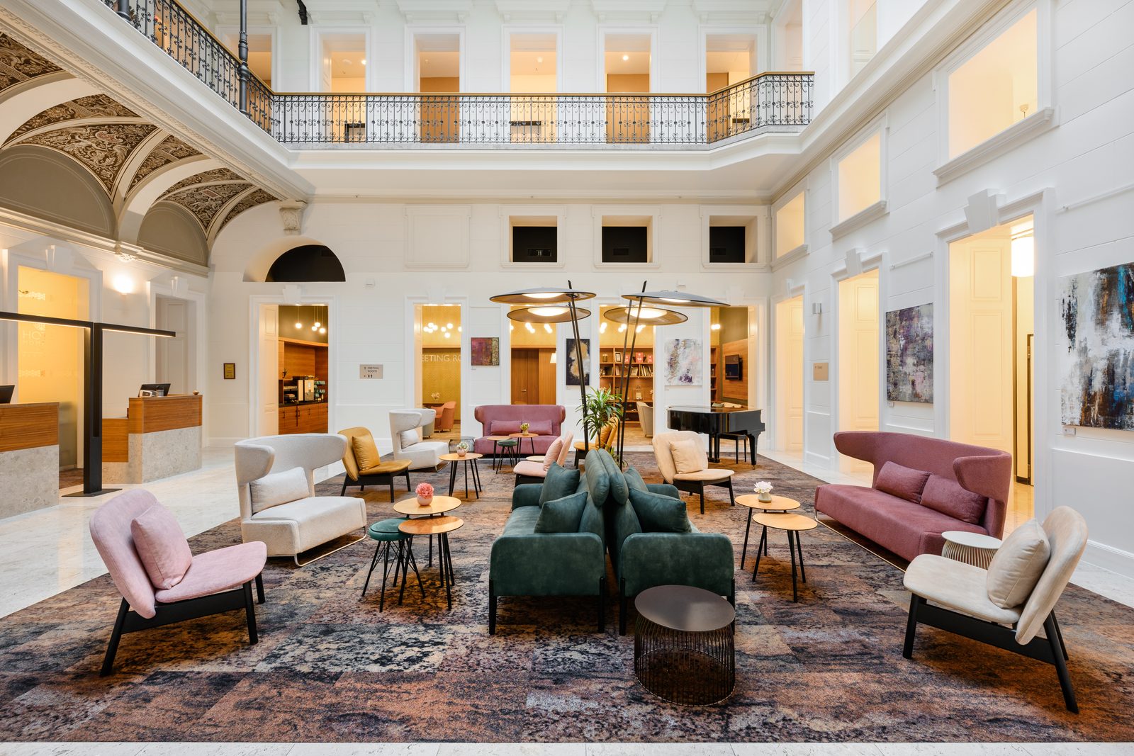 The white atrium lounge of Hotel Moments Budapest, with a wrought-iron gallery above, ornate painted vaults on the left, a grand piano and pink, green and cream armchairs and sofas on a large patterned rug