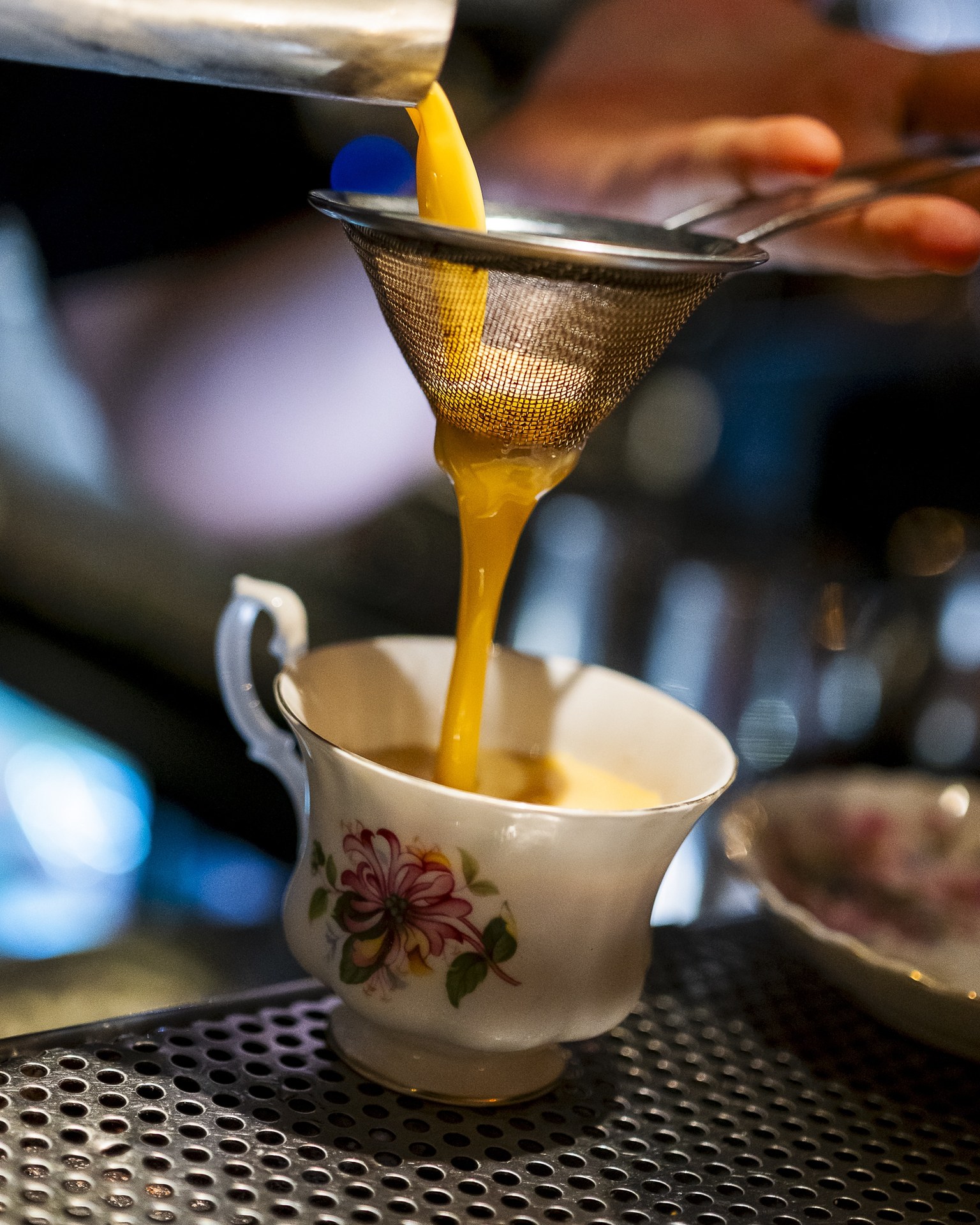 A yellow cocktail poured through a fine strainer into a floral porcelain teacup on the bar at Hotsy Totsy