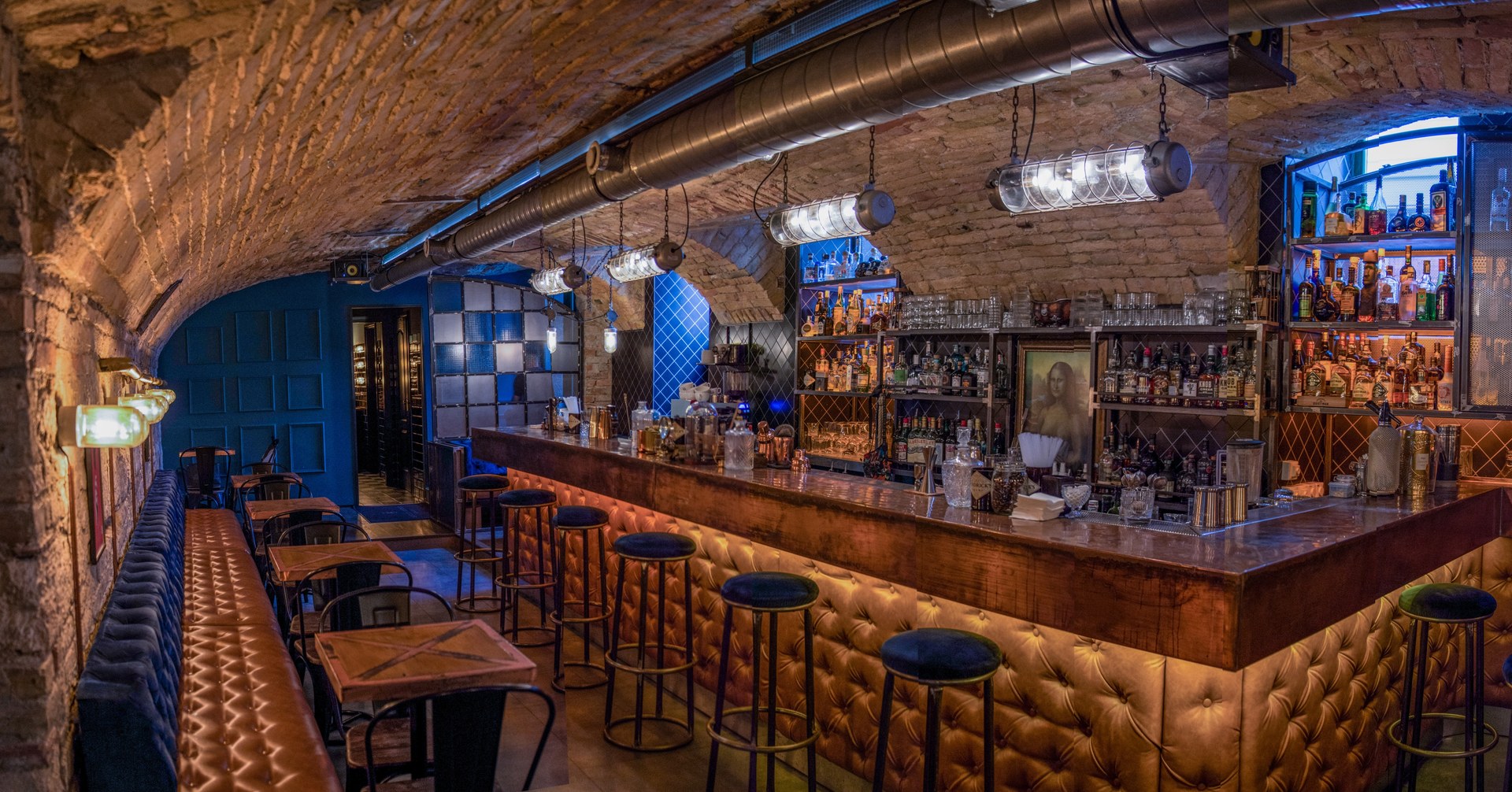 The bar at Hotsy Totsy under a vaulted brick ceiling, with a tufted leather counter, dark stools, backlit bottle shelves and blue-panelled walls