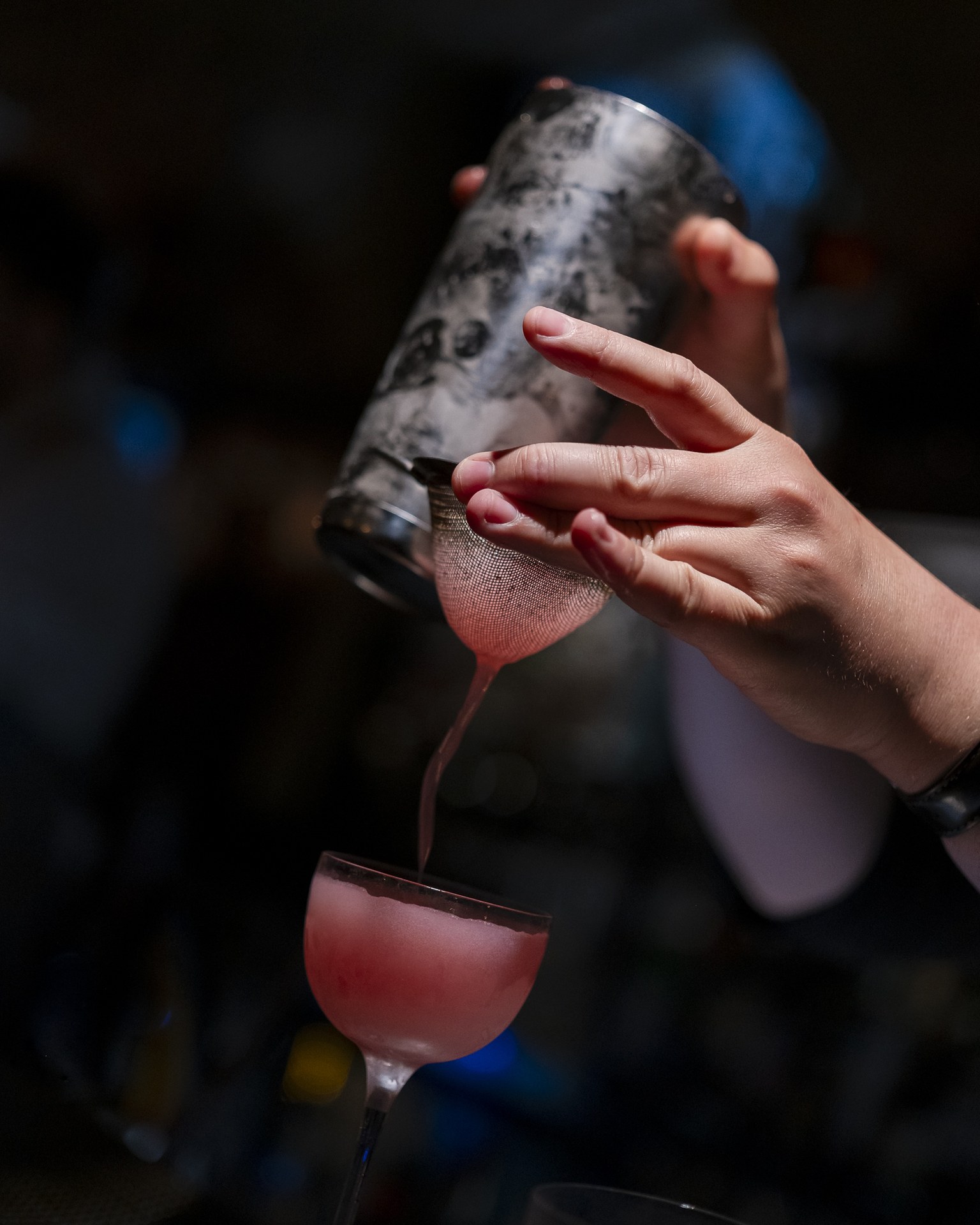 A hand at Hotsy Totsy fine-straining a pink cocktail from a metal shaker into a stemmed coupe