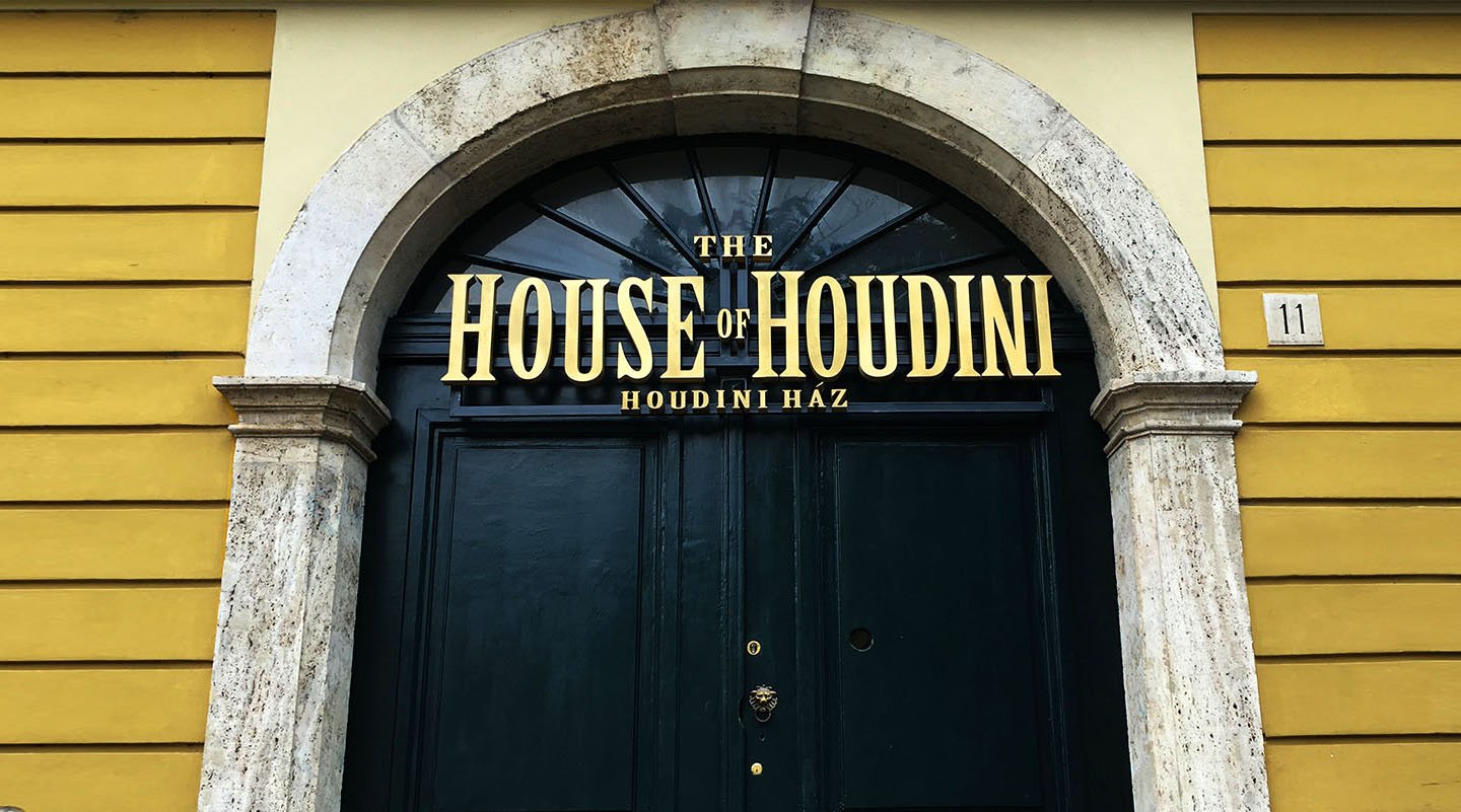 The arched dark green door of the House of Houdini at Dísz tér 11, with gold lettering reading The House of Houdini, Houdini Ház, on a yellow facade