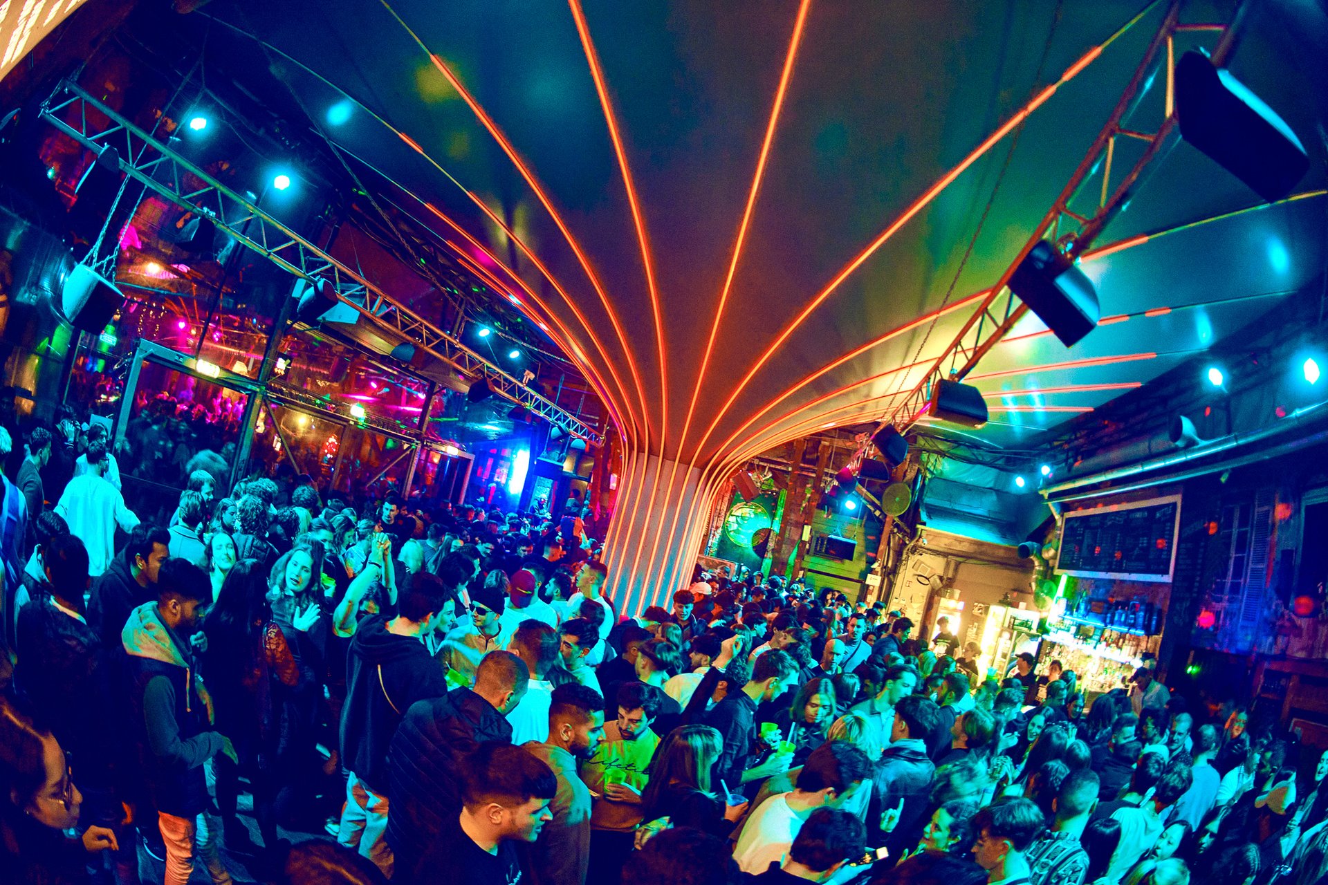 A packed courtyard at Instant-Fogas under a canopy of orange LED arcs, with the bar on the right