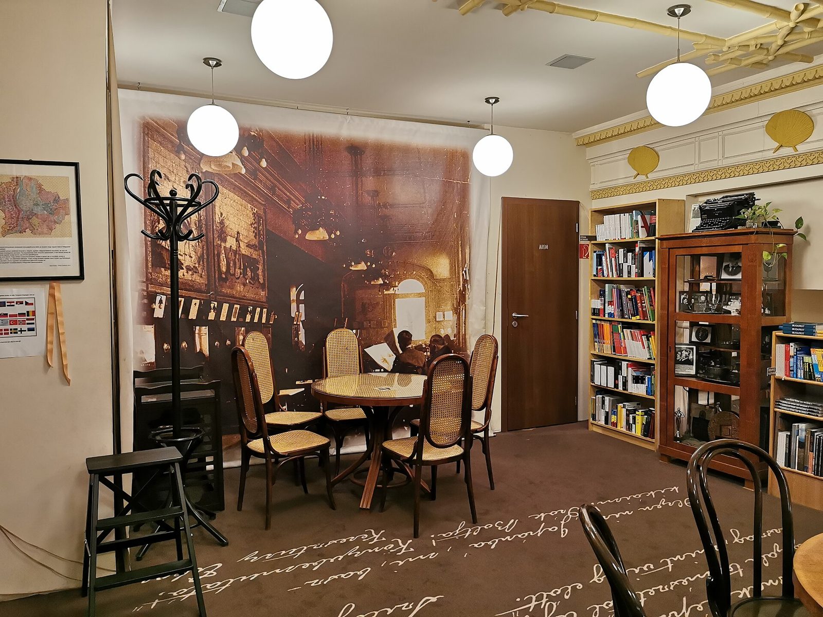 The tea room at the back of Írók Boltja, bentwood chairs around small round tables under globe lamps, bookshelves along the walls and a large sepia photograph of an old café hall on the far wall