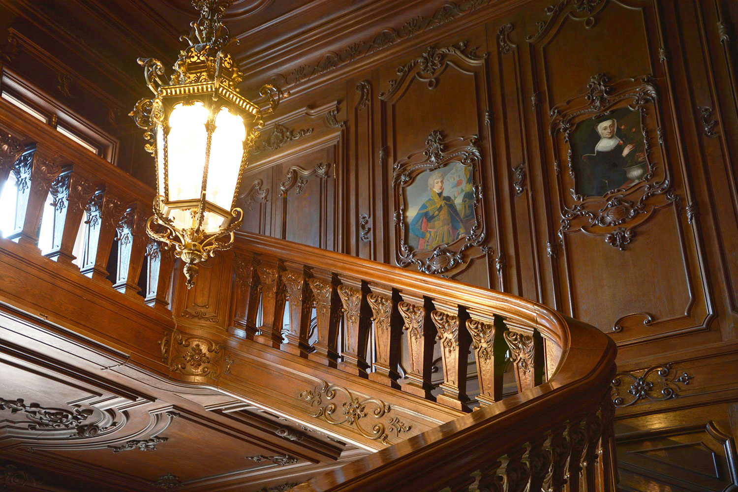 The carved oak staircase of the Festetics Palace, with a brass lantern, wood-paneled walls and family portraits