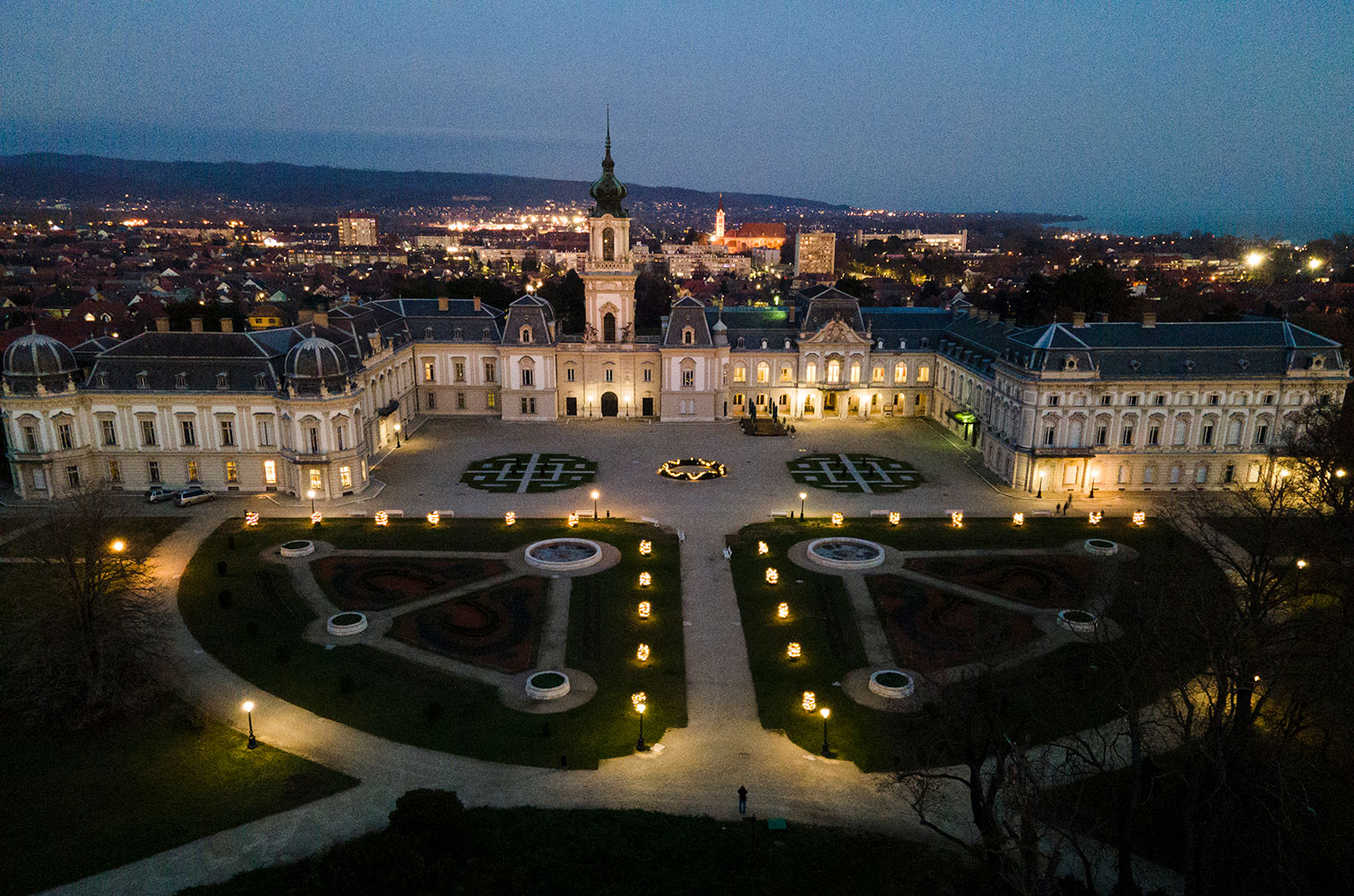 The Festetics Palace and its formal garden lit up at dusk, seen from above, with the town of Keszthely behind