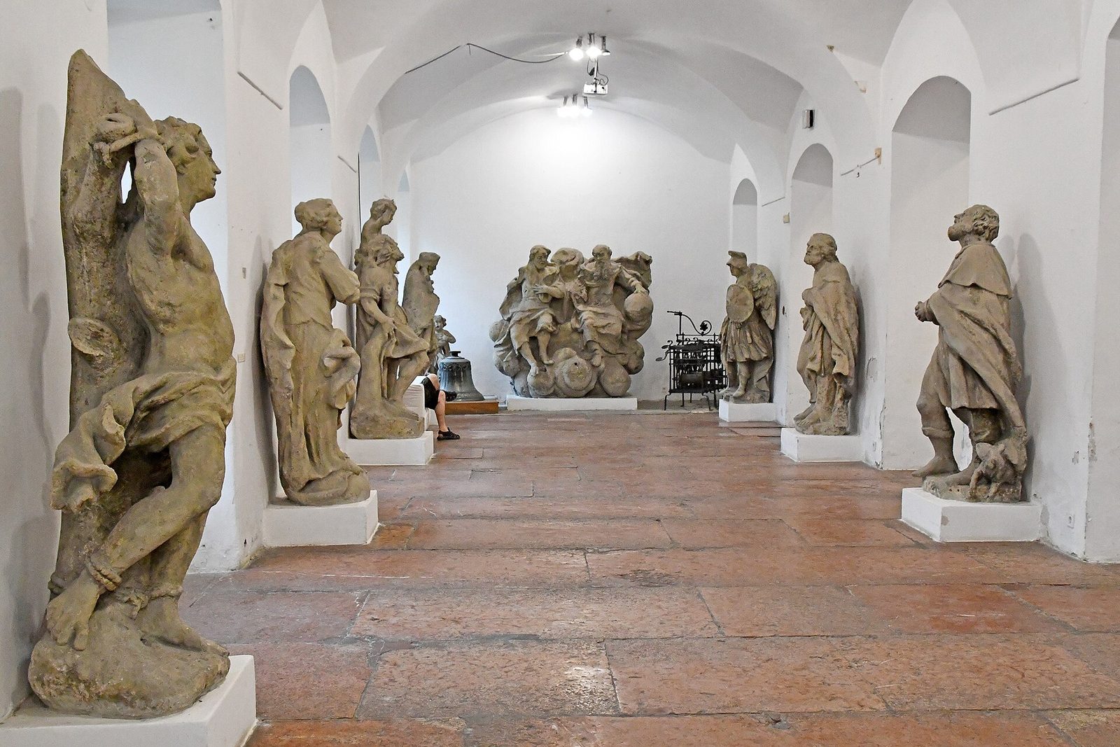 Baroque stone statues lining a long white vaulted hall in the Kiscelli Museum, with a large sculptural group at the far end