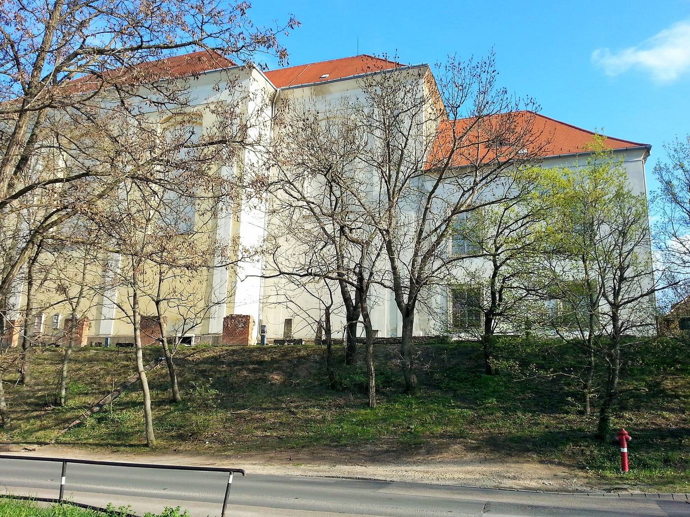 The long yellow former monastery of the Kiscelli Museum seen from the road below, behind bare trees on a grassy slope