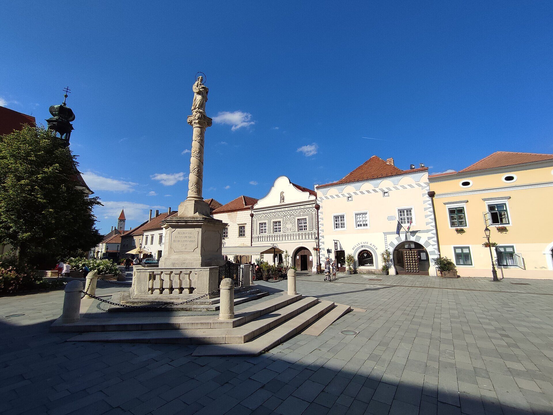 Jurisics tér v&nbsp;Kőszegu za slunečného dne, vysoký barokní sloup se sochou Panny Marie před pastelovými fasádami, jedna z&nbsp;nich se šedým sgrafitem