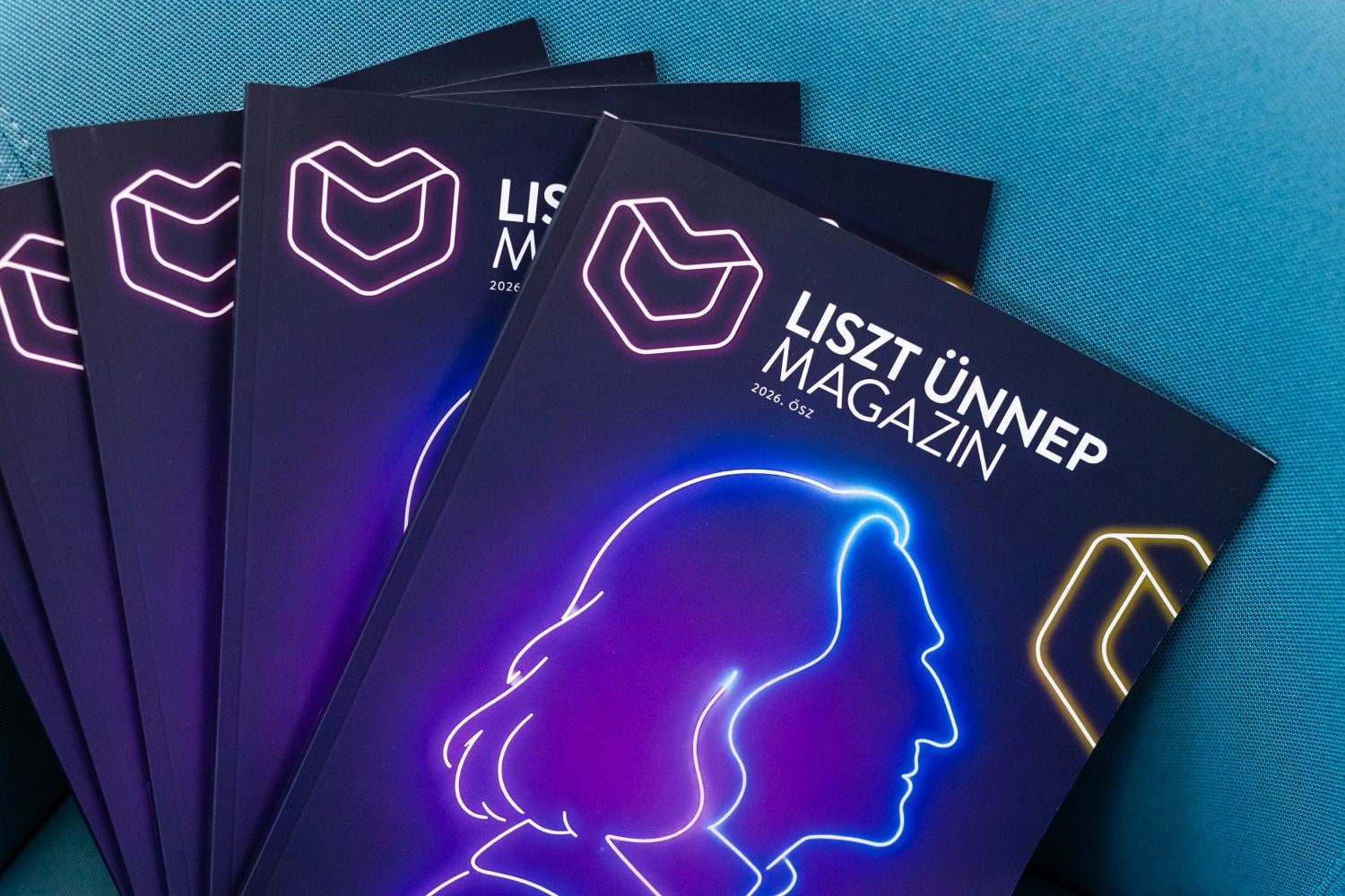 Copies of the Liszt Ünnep Magazin, autumn 2026 edition, fanned out on a blue armchair, each cover showing a neon-style outline of a long-haired profile