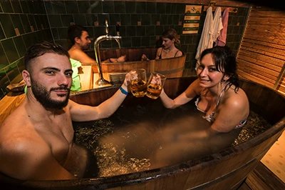 Two bathers raising mugs of beer in a wooden tub at the Lukács Beer Spa, with a second couple in another wooden tub and green tiles behind them