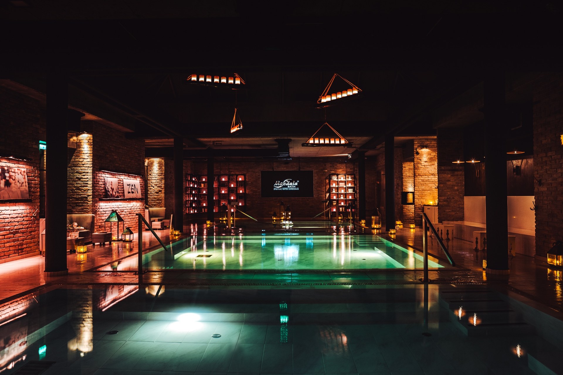 אולם הבריכות של Mandala Day Spa באור עמום, עם קירות לבנים חשופות, פנסים עם נרות לאורך השפה ובריכות מוארות עד מסך עם הלוגו של הספא