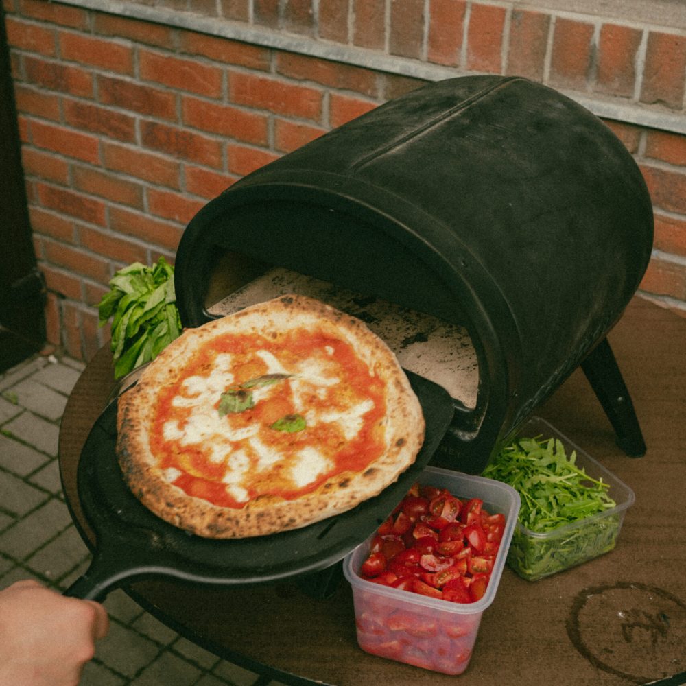 A Moto Margherita with a blistered, leopard-spotted crust on a peel in front of a small dome pizza oven, next to tubs of cherry tomatoes and arugula
