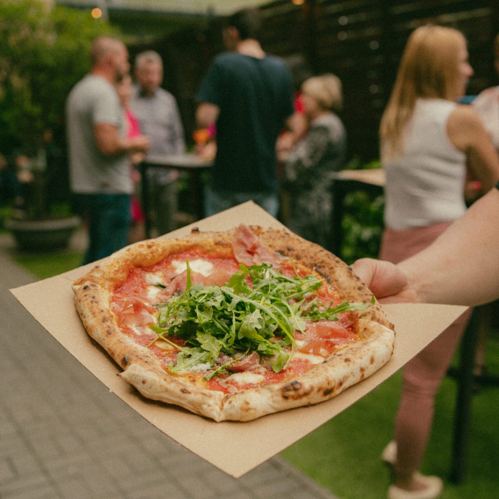 A Moto pizza topped with prosciutto and a pile of arugula, served on brown paper at an outdoor party