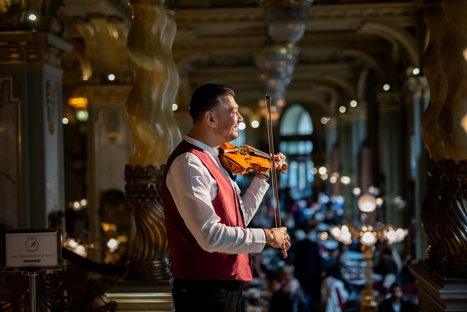 A violinist in a white shirt and red waistcoat playing on the gallery of New York Café, above the crowded café floor