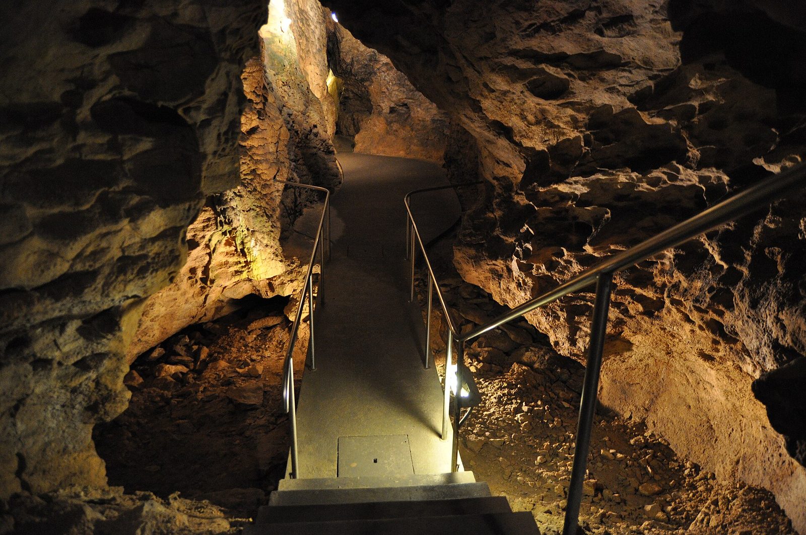 Steel stairs with handrails climbing through a lit chamber of the Pál-völgyi Cave, between yellow-brown limestone walls