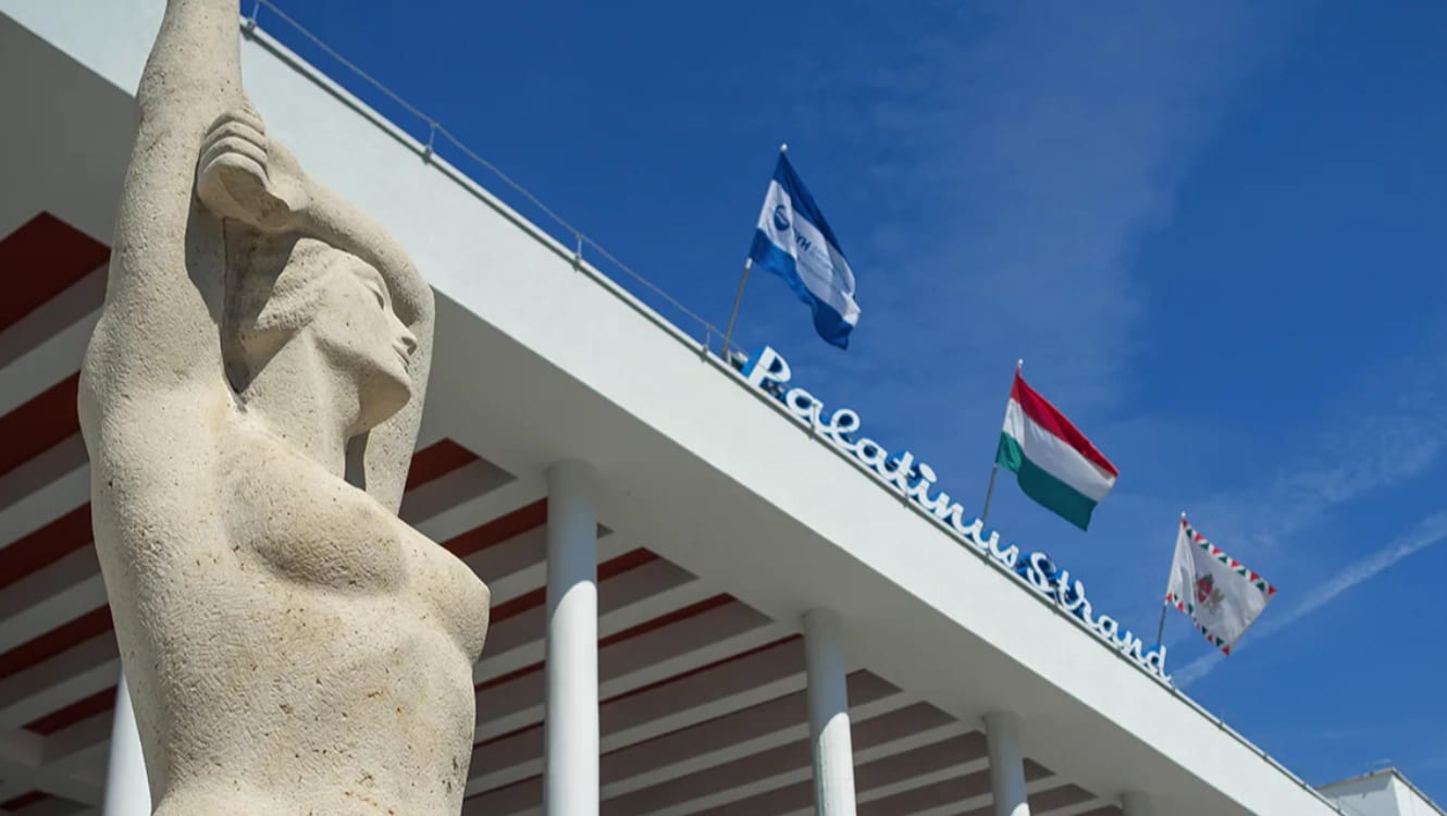 Statua in pietra di una donna con le braccia alzate davanti alla pensilina bianca dell’ingresso del Palatinus, con la scritta Palatinus Strand e tre bandiere contro il cielo blu