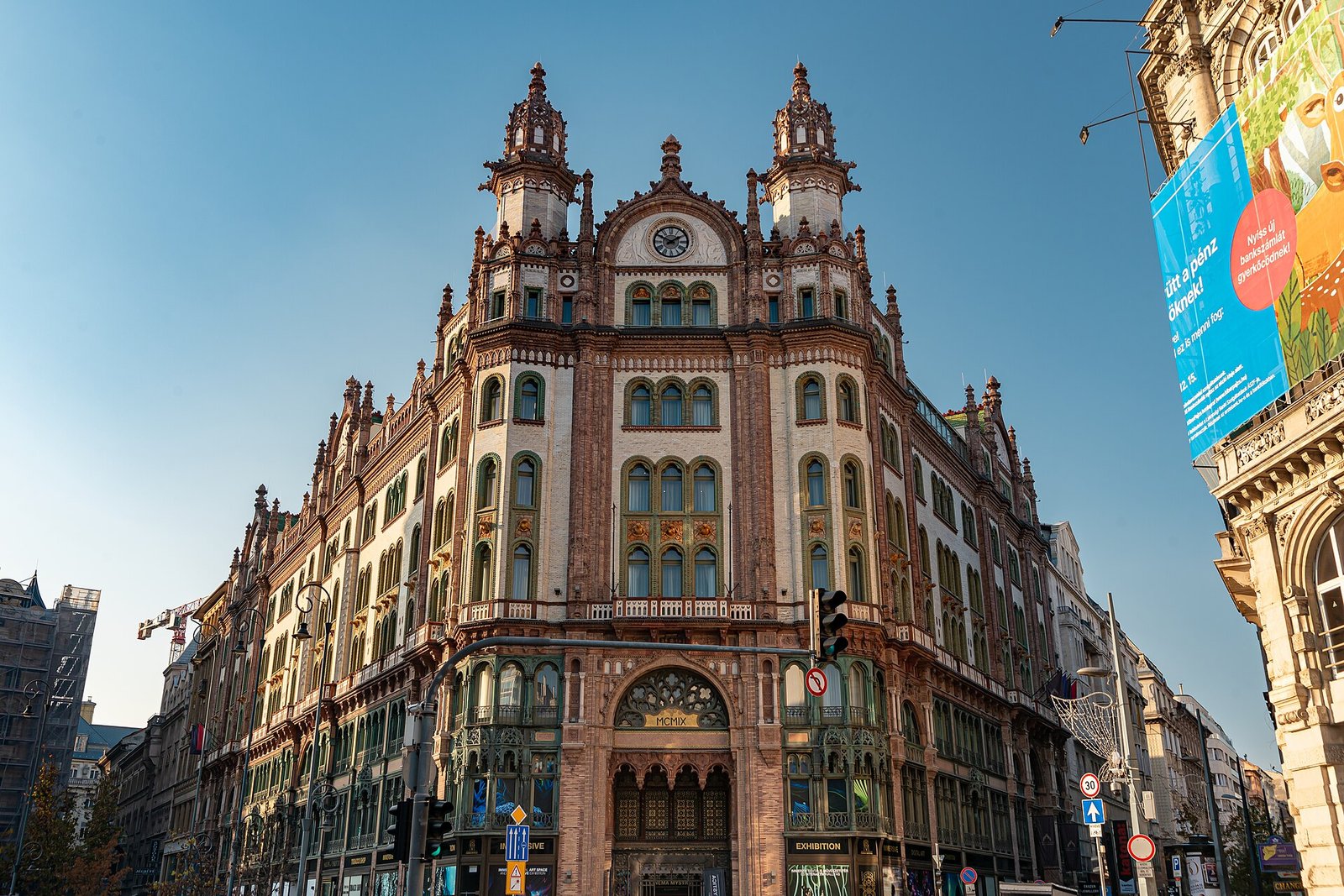 The corner facade of the Párisi Udvar on Ferenciek tere in Budapest: richly ornamented brick and ceramic stories under two pinnacled towers and a clock, with an arched entrance at street level