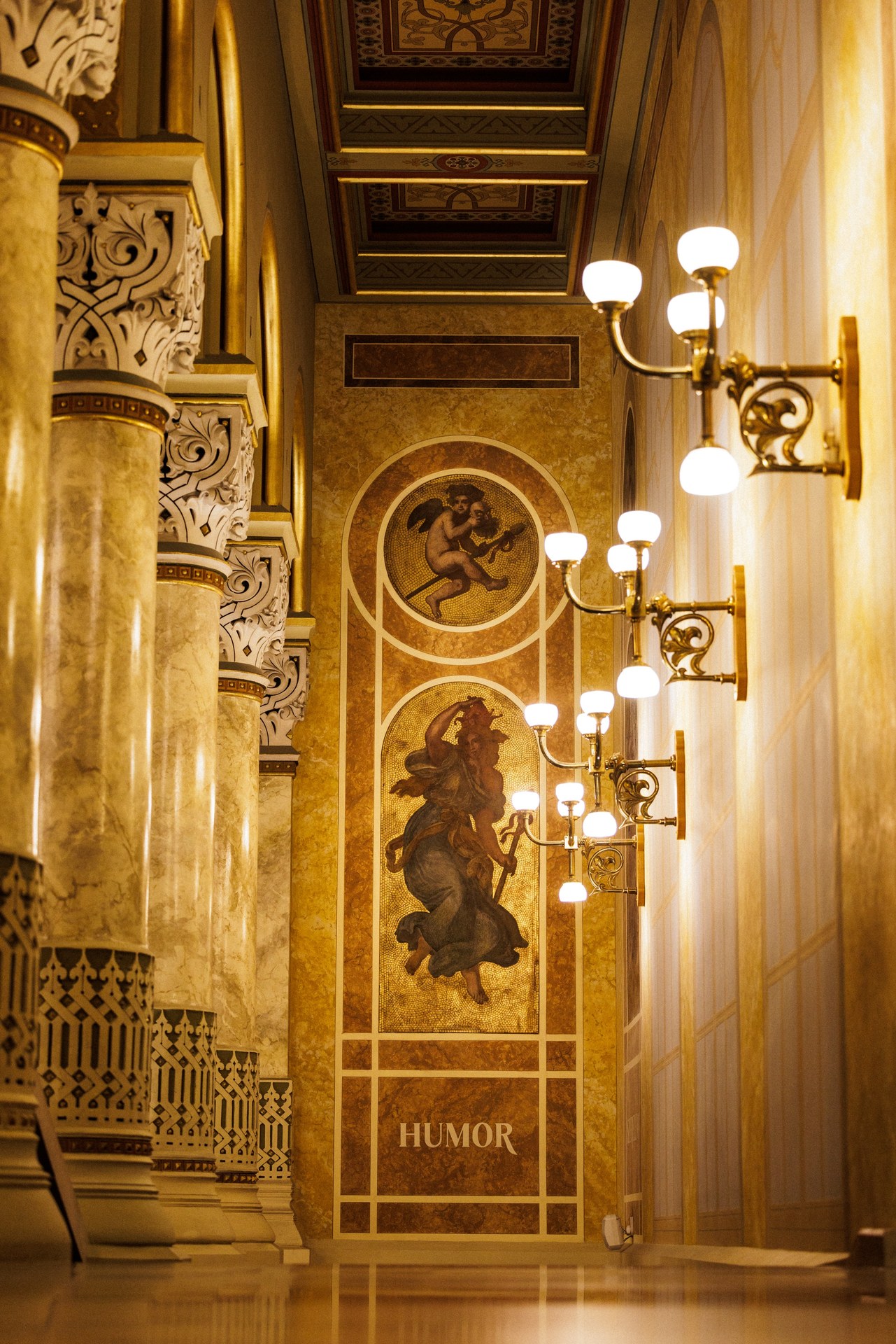 A corridor inside the Pesti Vigadó with marble-effect columns, brass wall lamps and a mosaic panel of a robed figure above the word HUMOR