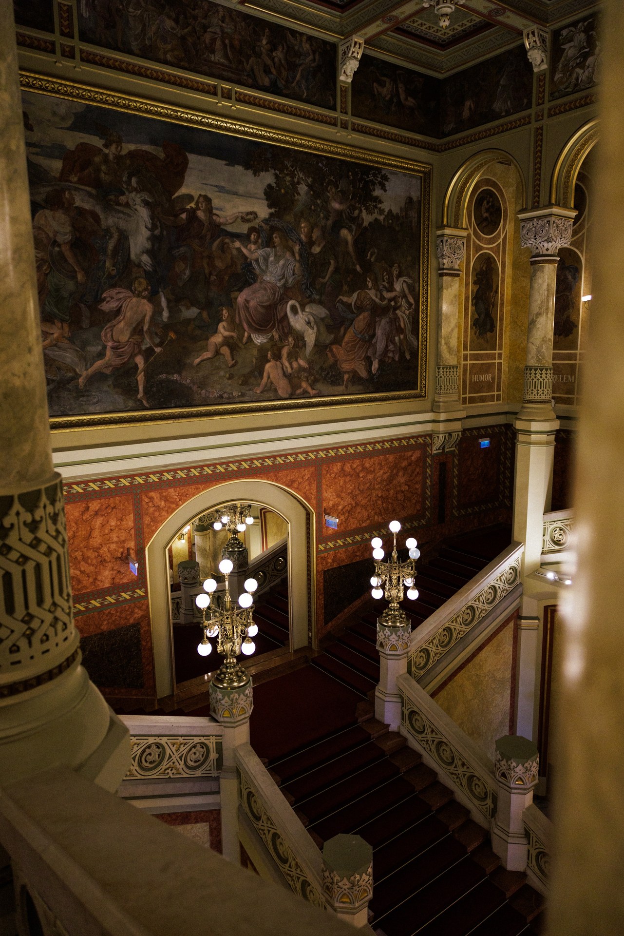 The Grand Staircase of the Pesti Vigadó seen from an upper landing, with red-carpeted steps, brass lamp standards and a large painted canvas on the wall above