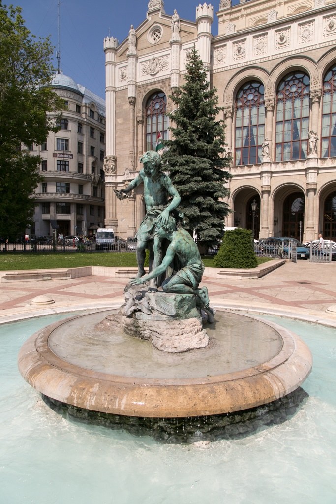A bronze fountain of two children in front of the Pesti Vigadó, with the building’s arched windows and carved facade behind a fir tree