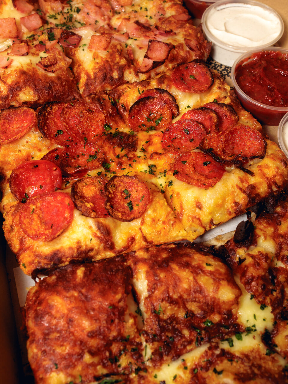 Square Pizzaművek pan pizzas topped with salami, with browned cheesy edges, next to cups of sour-cream and tomato dip