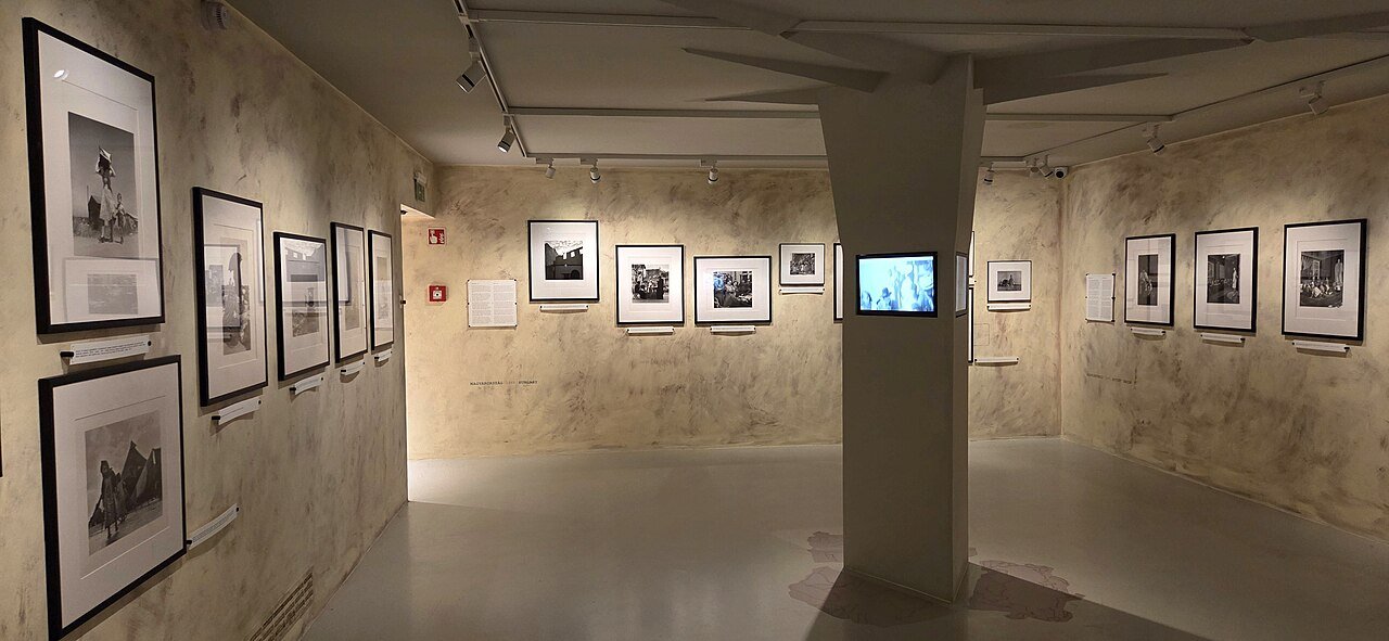 Další sál Capovy výstavy se zarámovanými černobílými fotografiemi na třech stěnách a malou obrazovkou zabudovanou do bílého sloupu