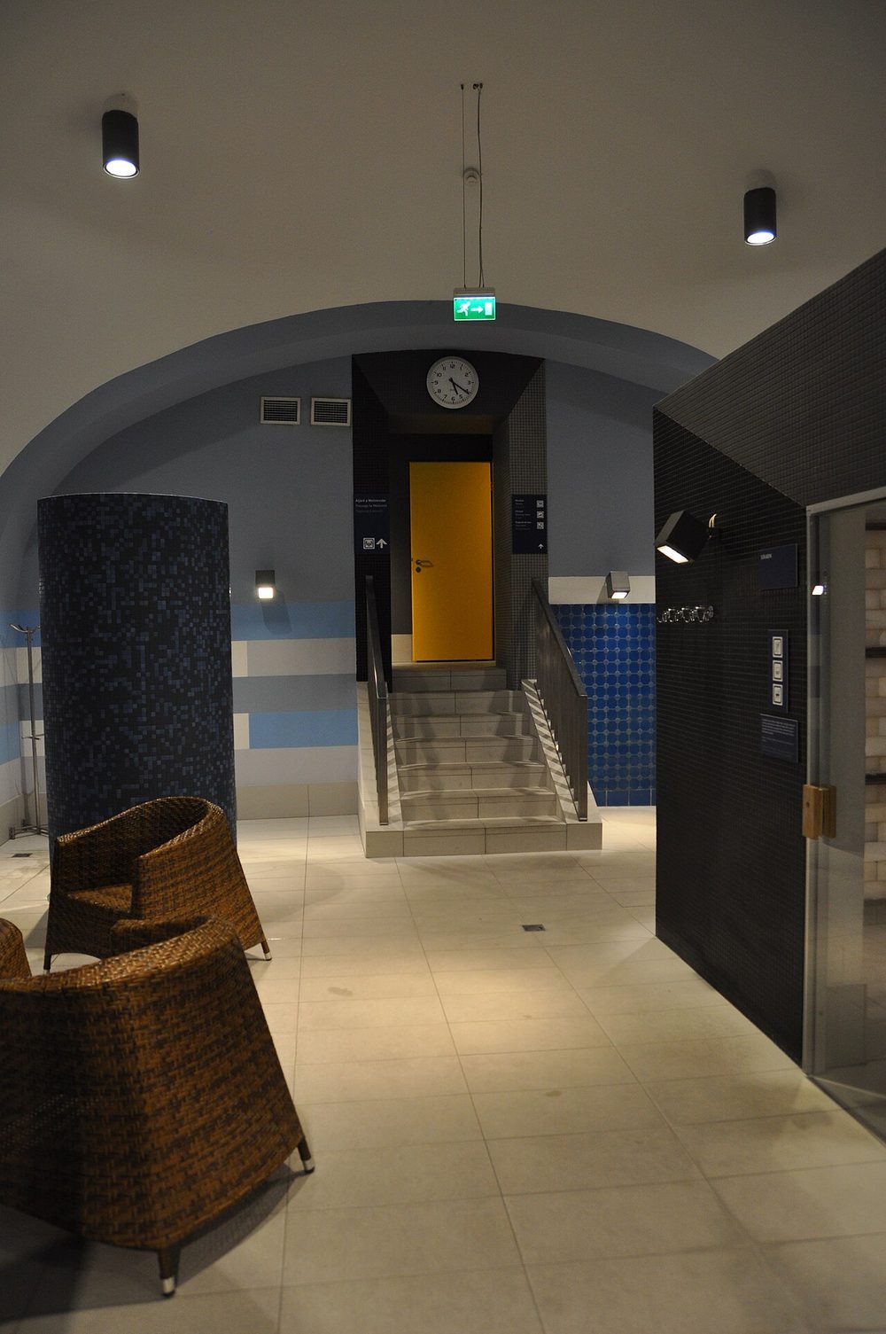 Le coin repos de l’espace sauna des bains Rudas, avec deux fauteuils en osier sur un sol carrelé, une colonne en mosaïque et un petit escalier menant à une porte jaune