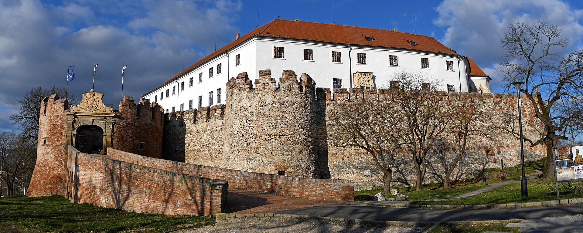 Bílé palácové křídlo hradu Siklós s&nbsp;červenou taškovou střechou nad cimbuřím kamenných hradeb, kulatá bašta a&nbsp;cihlová brána s&nbsp;barokním erbem