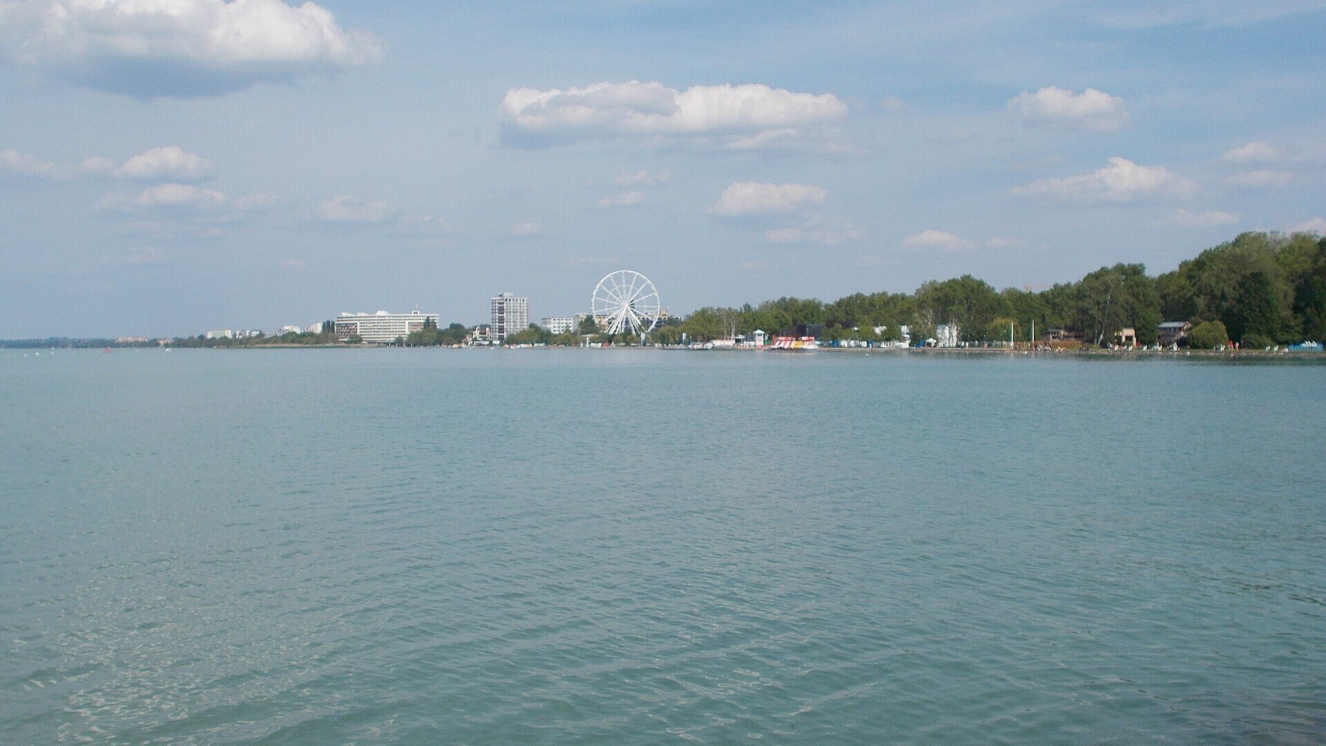 Lake Balaton seen from Siófok pier, looking across calm turquoise water toward the Nagystrand shoreline with a Ferris wheel and hotel towers