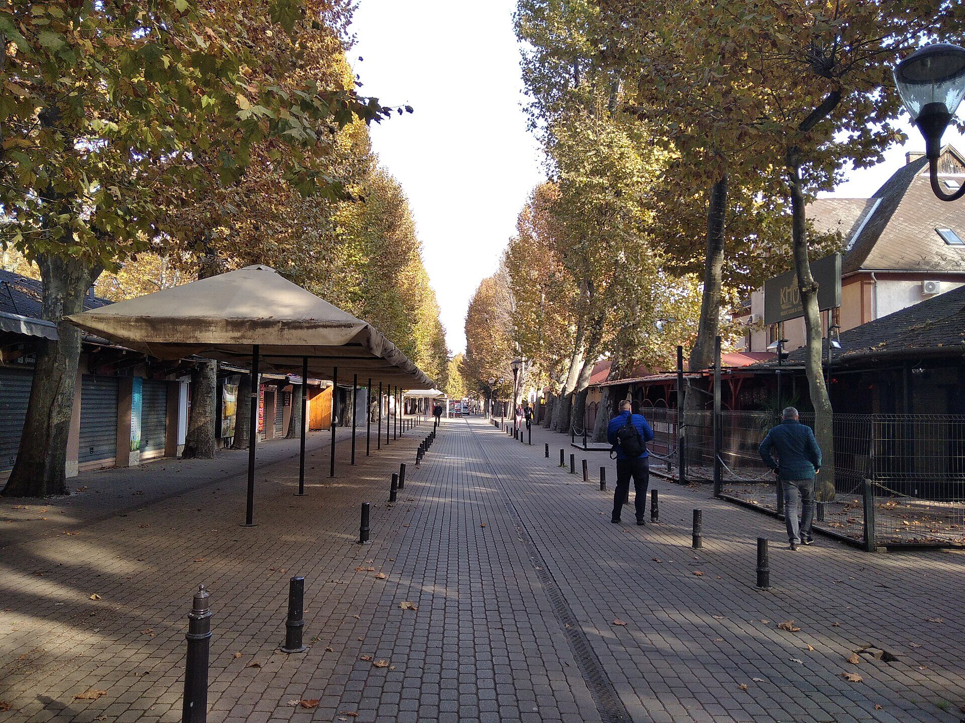 Petőfi promenade in Siófok in autumn, a paved tree-lined walk with shuttered kiosks and a few people walking under yellowing plane trees