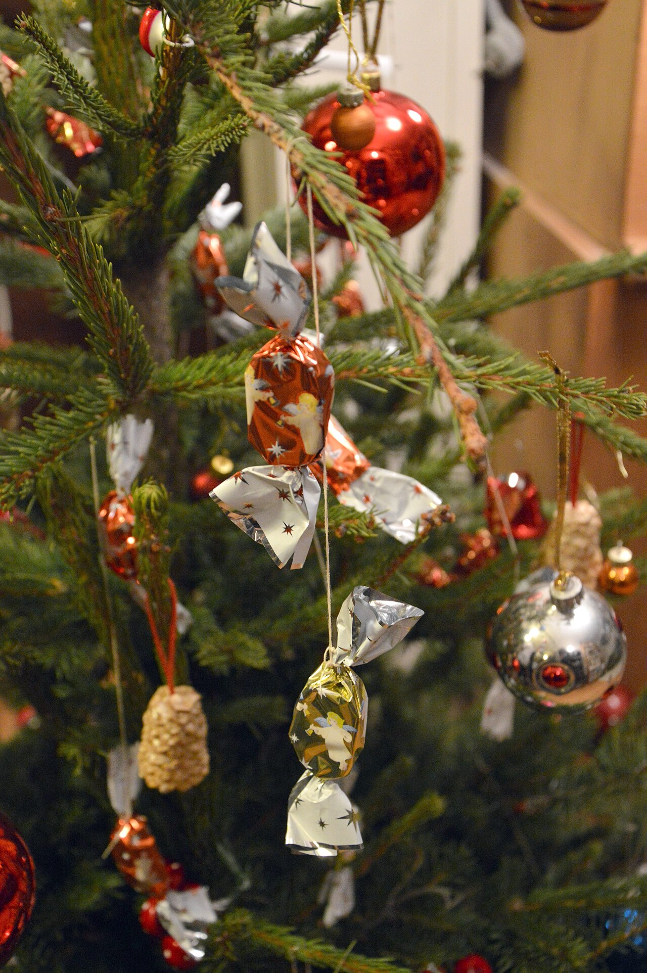 Szaloncukor sweets in red, gold and silver foil with angel prints, hanging from a Christmas tree beside glass baubles