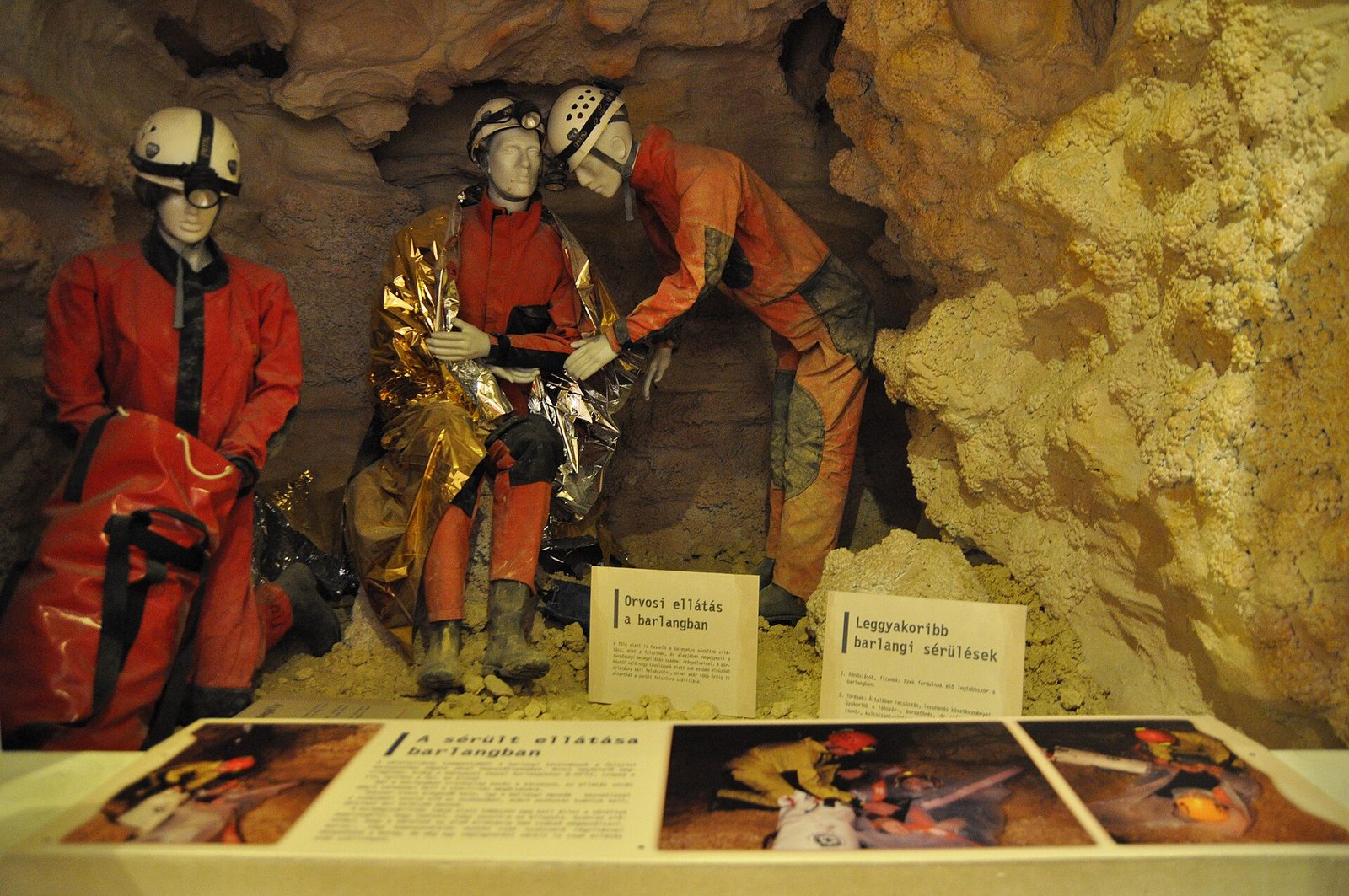 Caver mannequins in red overalls, helmets and headlamps tending an injured caver wrapped in a foil blanket, in the cave rescue display of the Szemlő-hegyi visitor center
